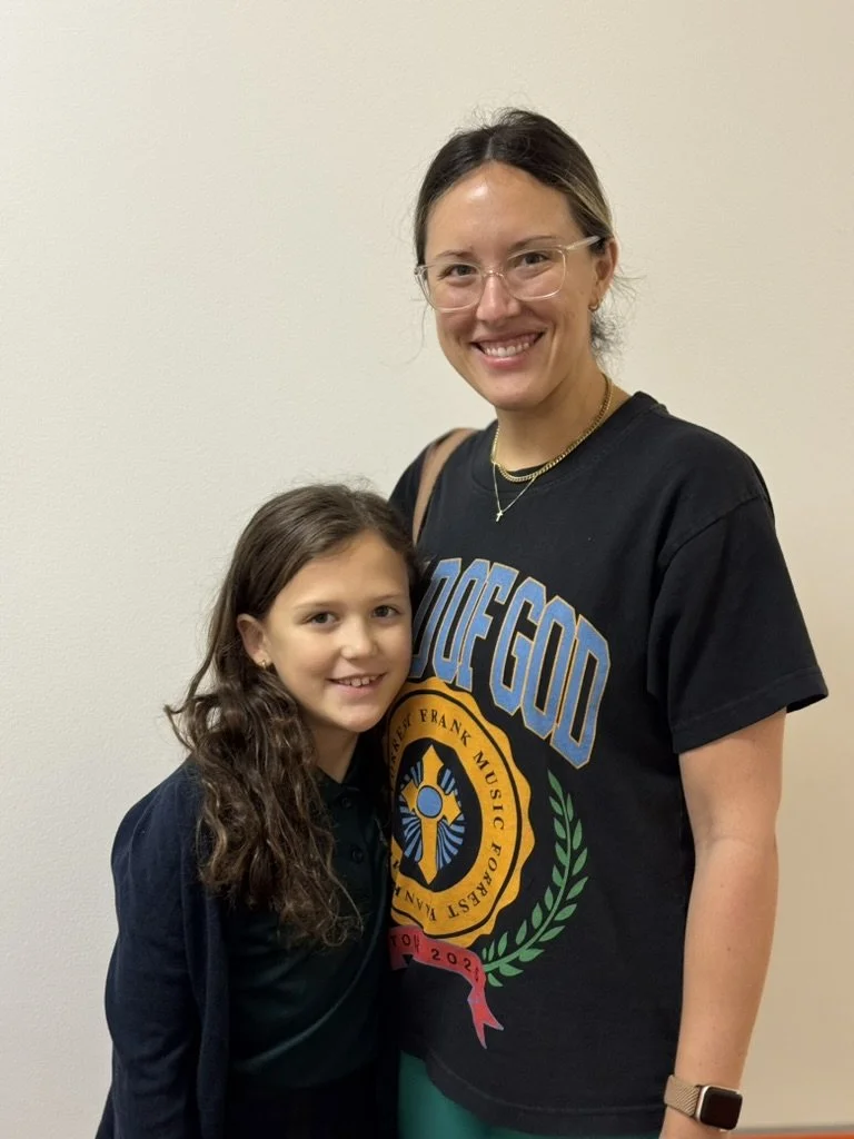 A smiling woman and a young girl posing together indoors against a plain wall.