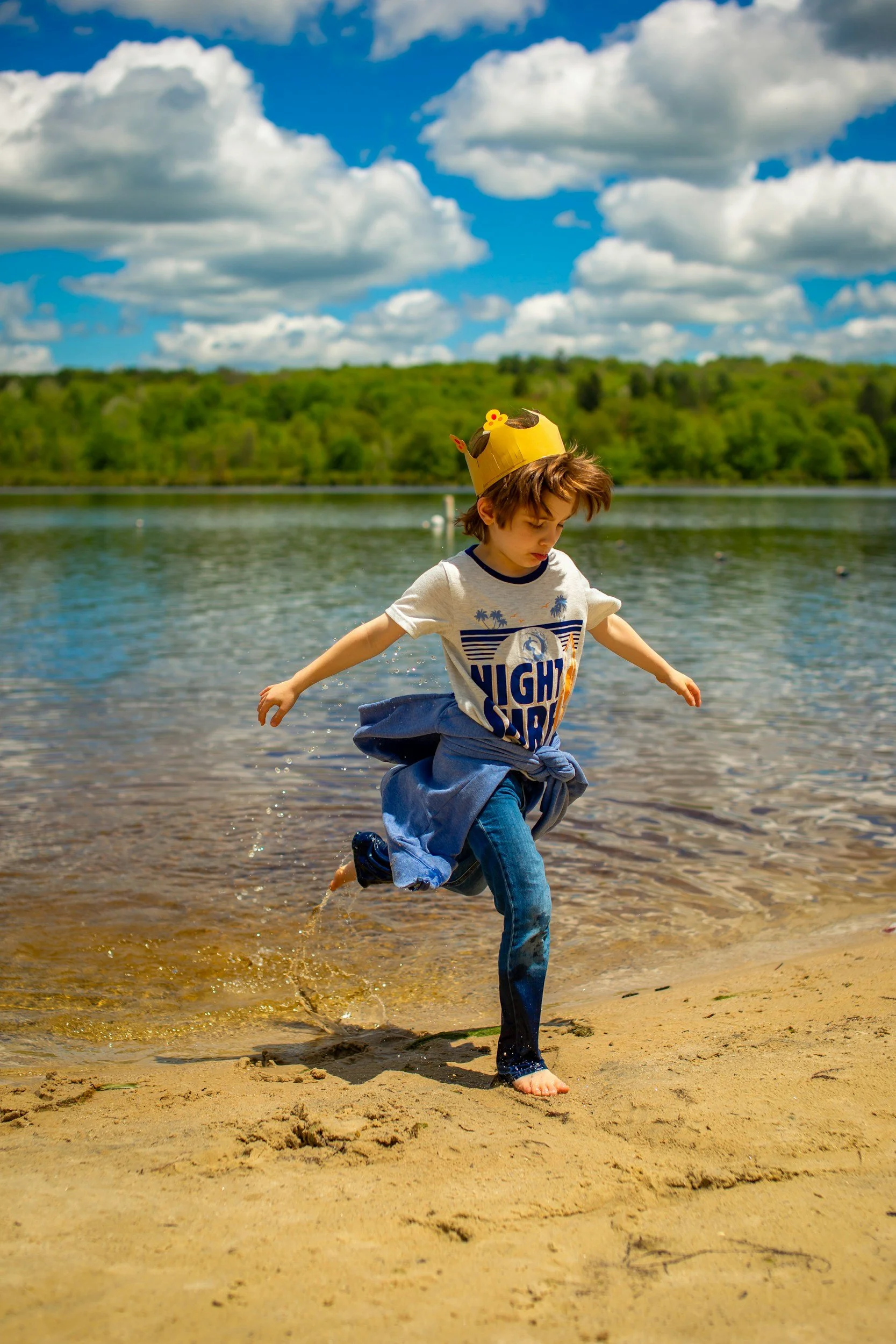 A young boy wearing a paper crown and a "Night Surf" t-shirt runs along a sandy beach near a body of water, with a forested area in the background under a partly cloudy sky.
