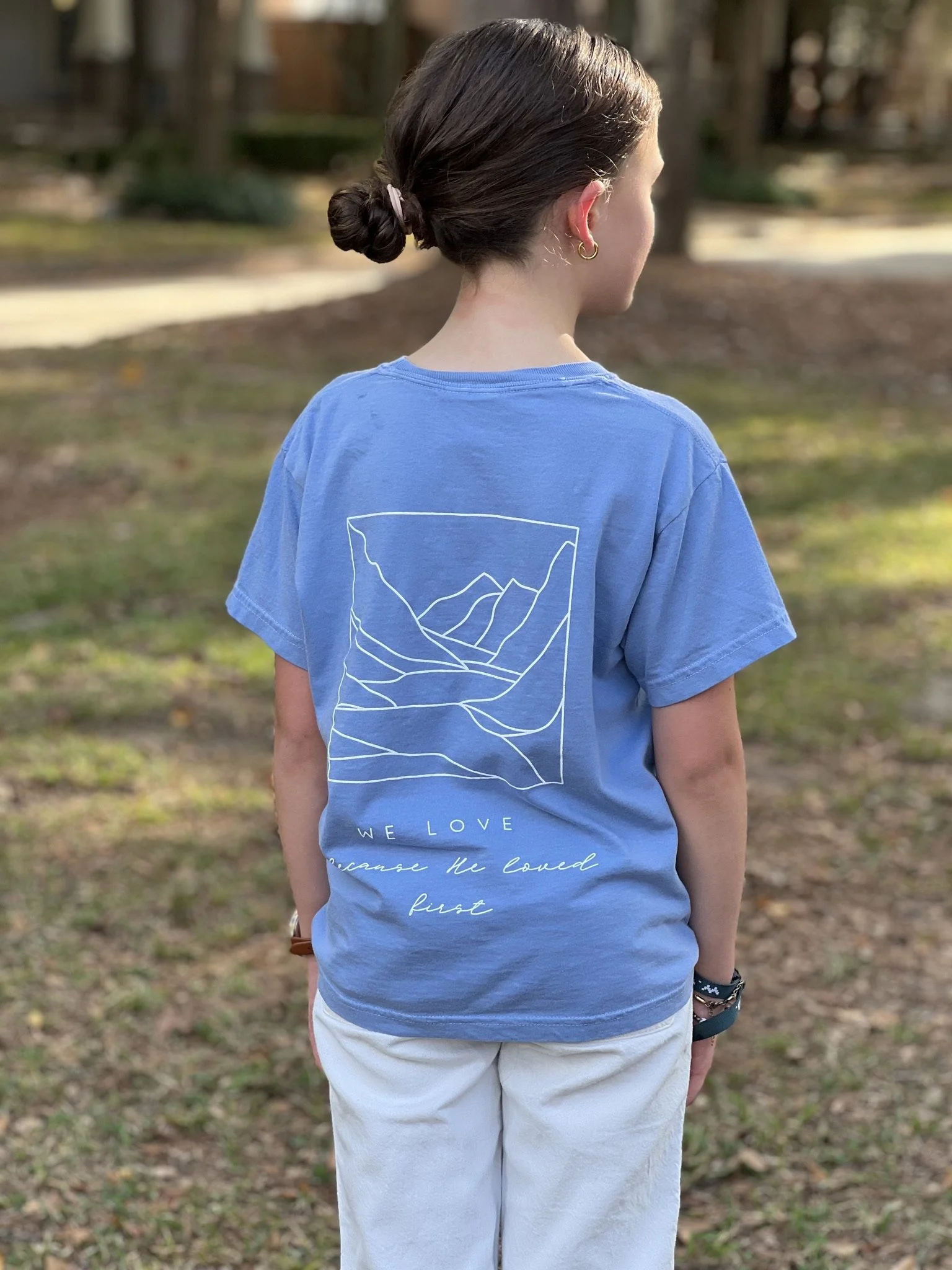 Back of a girl wearing a blue T-shirt with a mountain line art design and the text 'We love because he loved first,' standing outdoors.