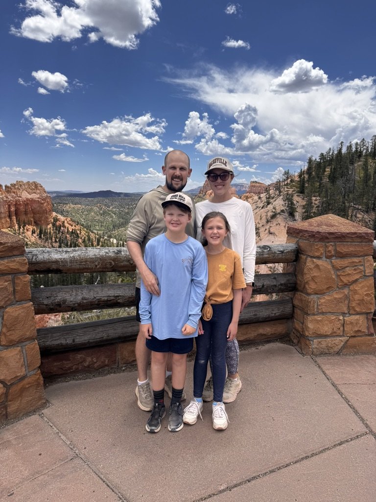 Family of four posing at a scenic overlook with red rock formations, trees, and a cloudy blue sky in the background.