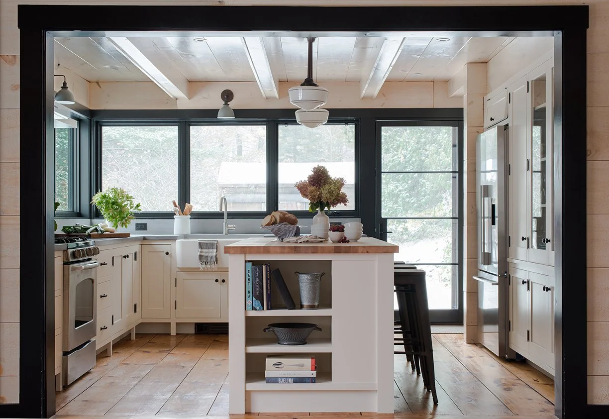 Bright, modern kitchen with white cabinets, wooden countertops, large windows, and a central island with shelves. Decor includes a vase of flowers, books, and bowls. Sliding glass door leads outside.