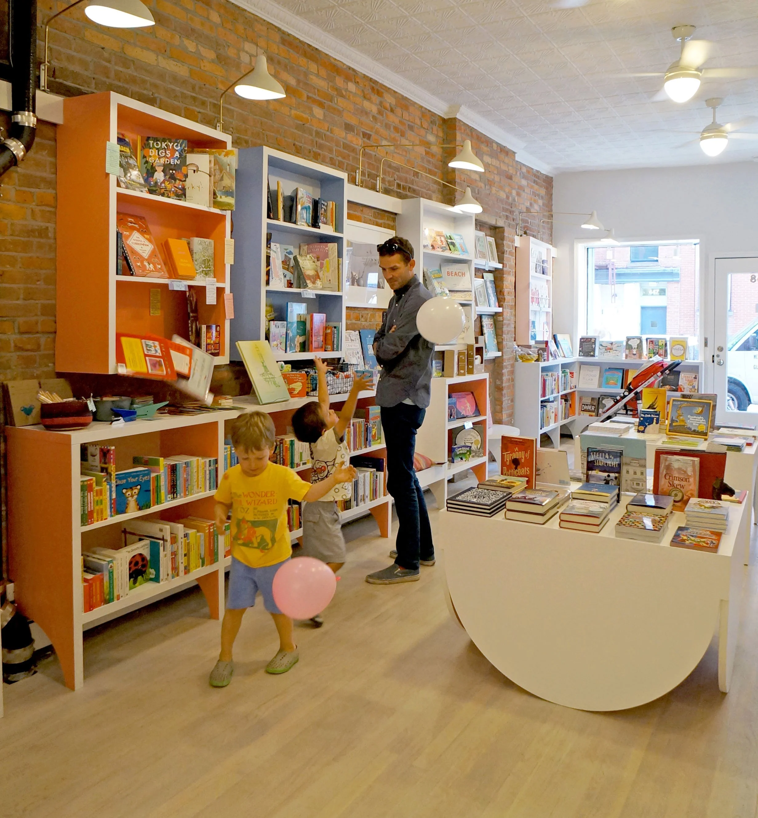 Inside a cozy bookstore with brick walls and white shelves filled with children's books. Two young boys are playing with balloons while an adult man stands nearby. Bright lighting and large windows let in natural light.