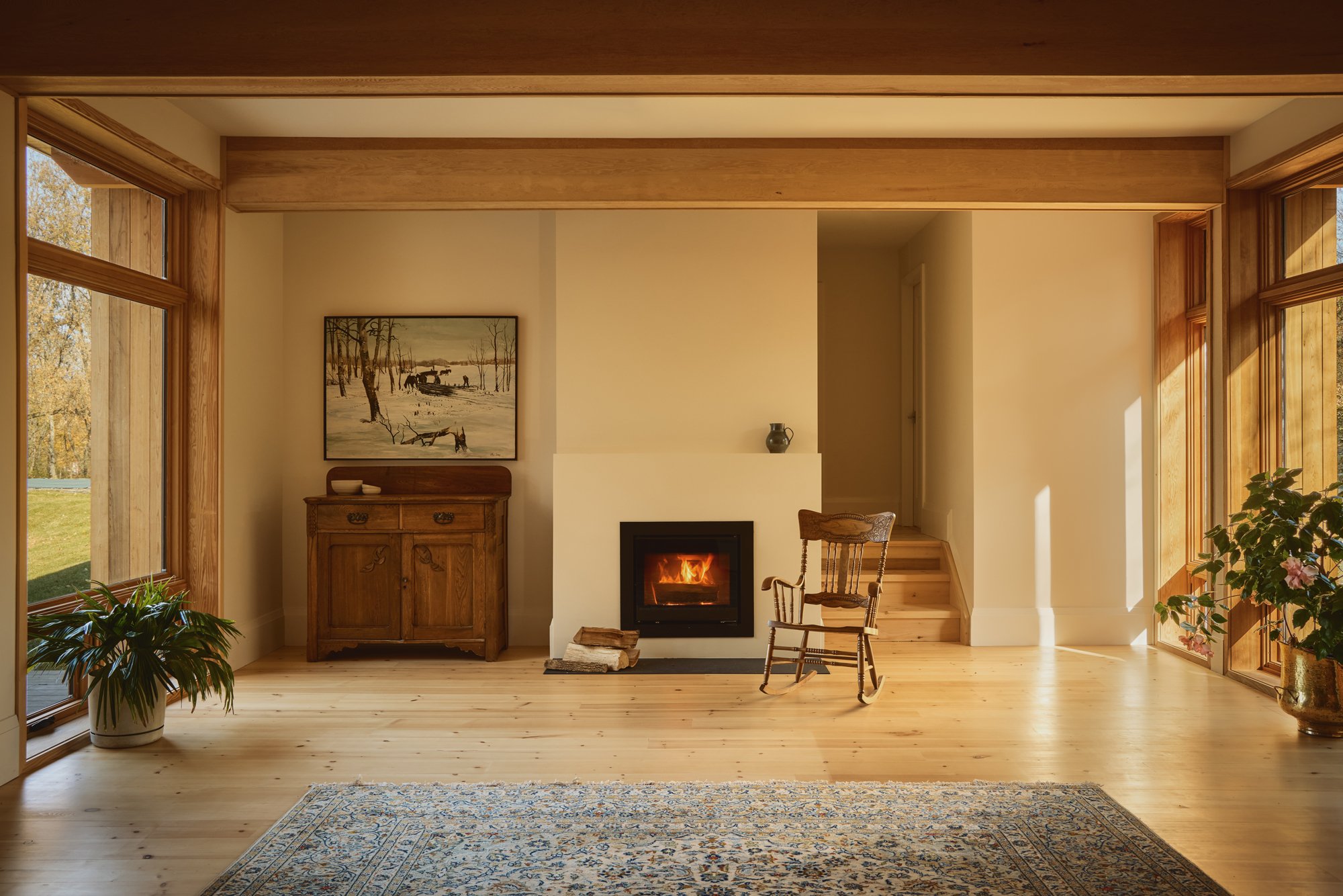 Living room with a fireplace, wooden furniture, and large windows letting in sunlight, featuring plants and a patterned rug.