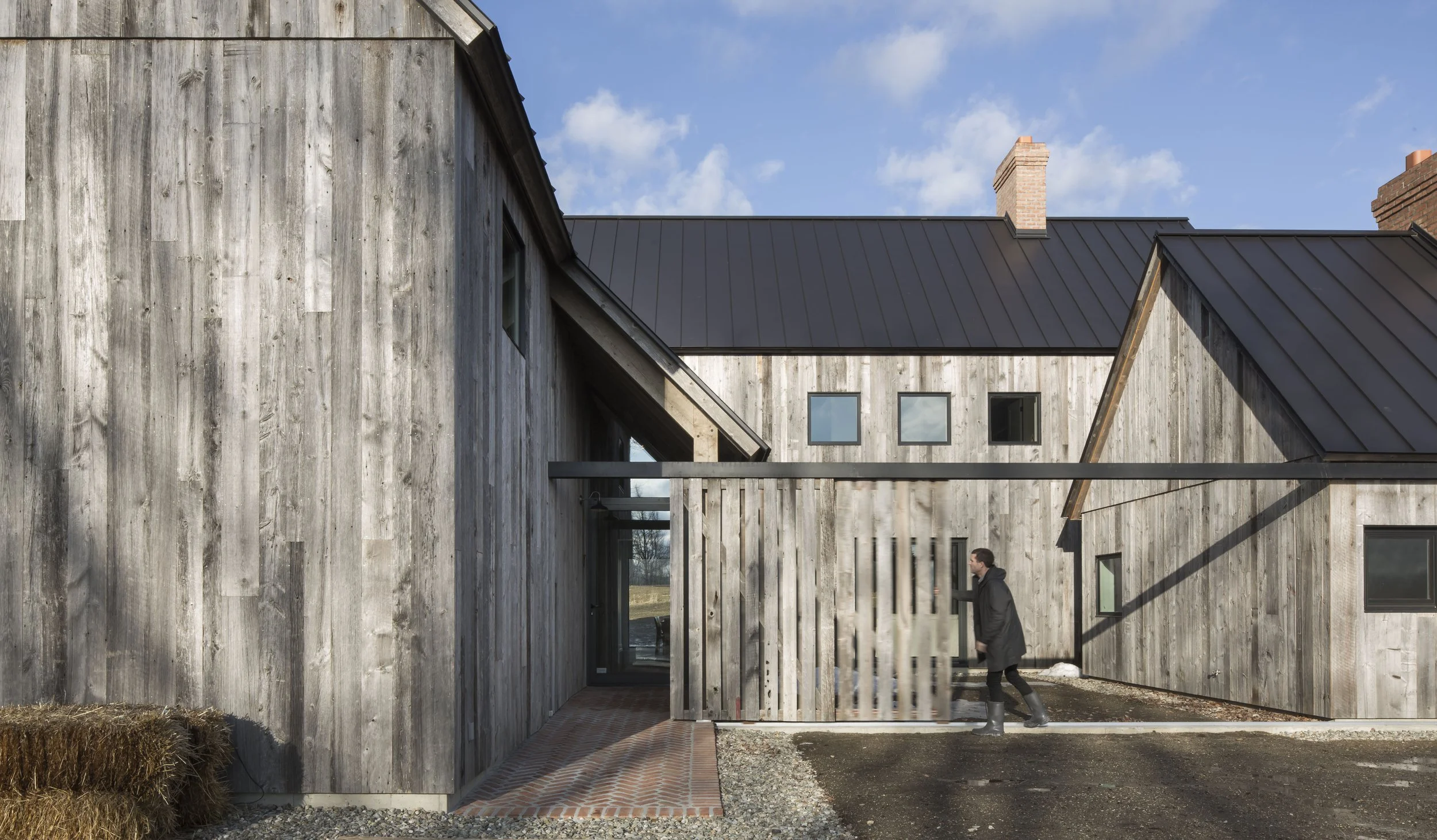 A person walking past a modern building with weathered wooden siding and multiple windows, under a partly cloudy sky.