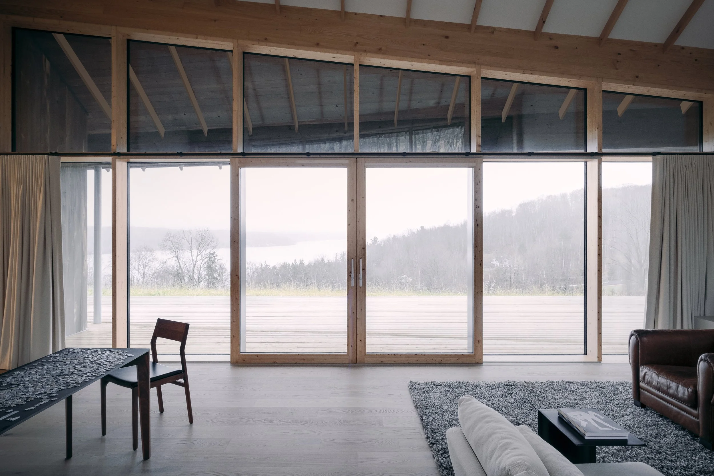 Living room with large sliding glass doors and windows overlooking mountains and a lake in the distance; interior features hardwood floors, a gray rug, a white sofa, a dark leather armchair, a black coffee table with a magazine, and a wooden dining table with a chair, with curtains on the side.