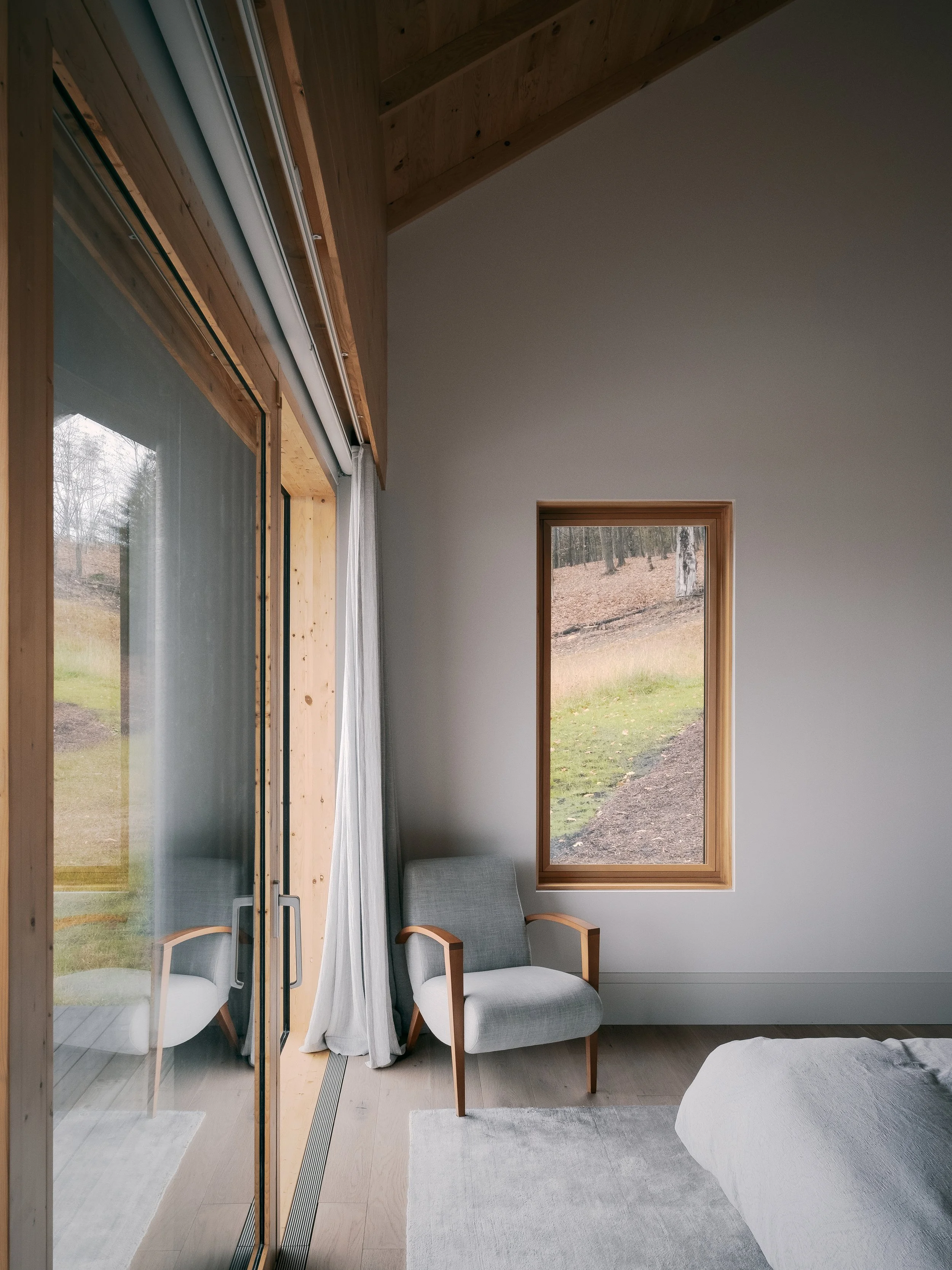 A bedroom corner with a wooden-framed window showing an outdoor landscape, a gray armchair with wooden armrests, a corner of a bed with white bedding, and a sliding glass door with sheer curtains leading to a small balcony or patio.