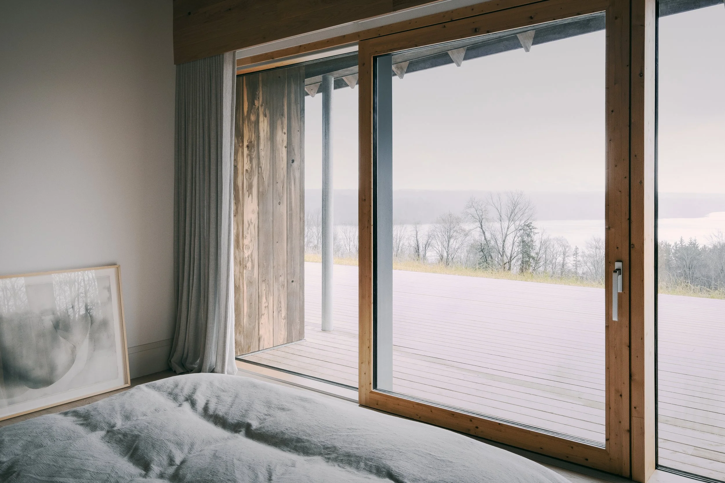 Bedroom with large glass sliding door opening to a wooden deck and a view of trees and water in the distance.