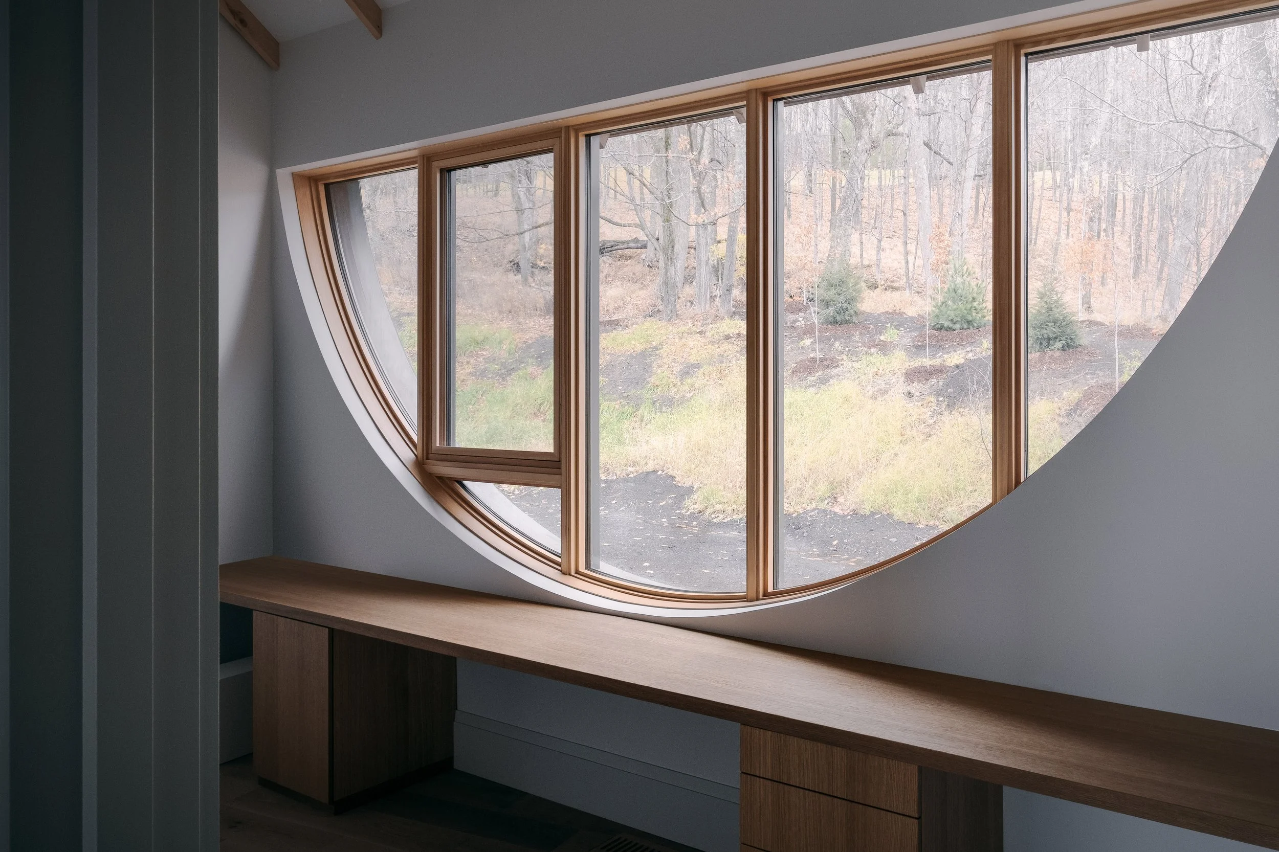 Interior of a room with a large, uniquely shaped window resembling an arc and a moon, with a view of a wooded hillside outside.