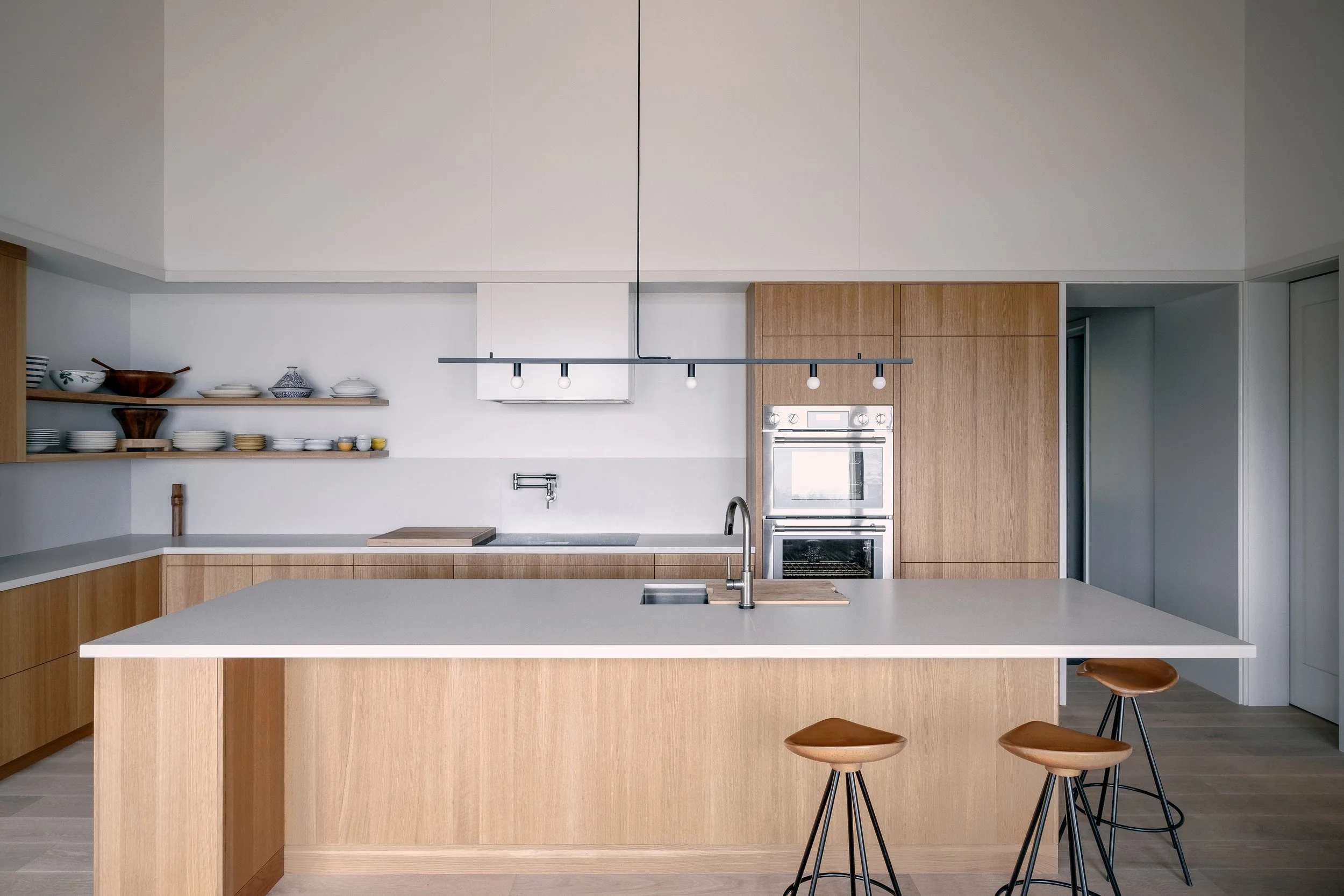 Modern kitchen with wooden cabinets, white countertops, open shelving with dishes, built-in oven, island with seating, and minimal decor.