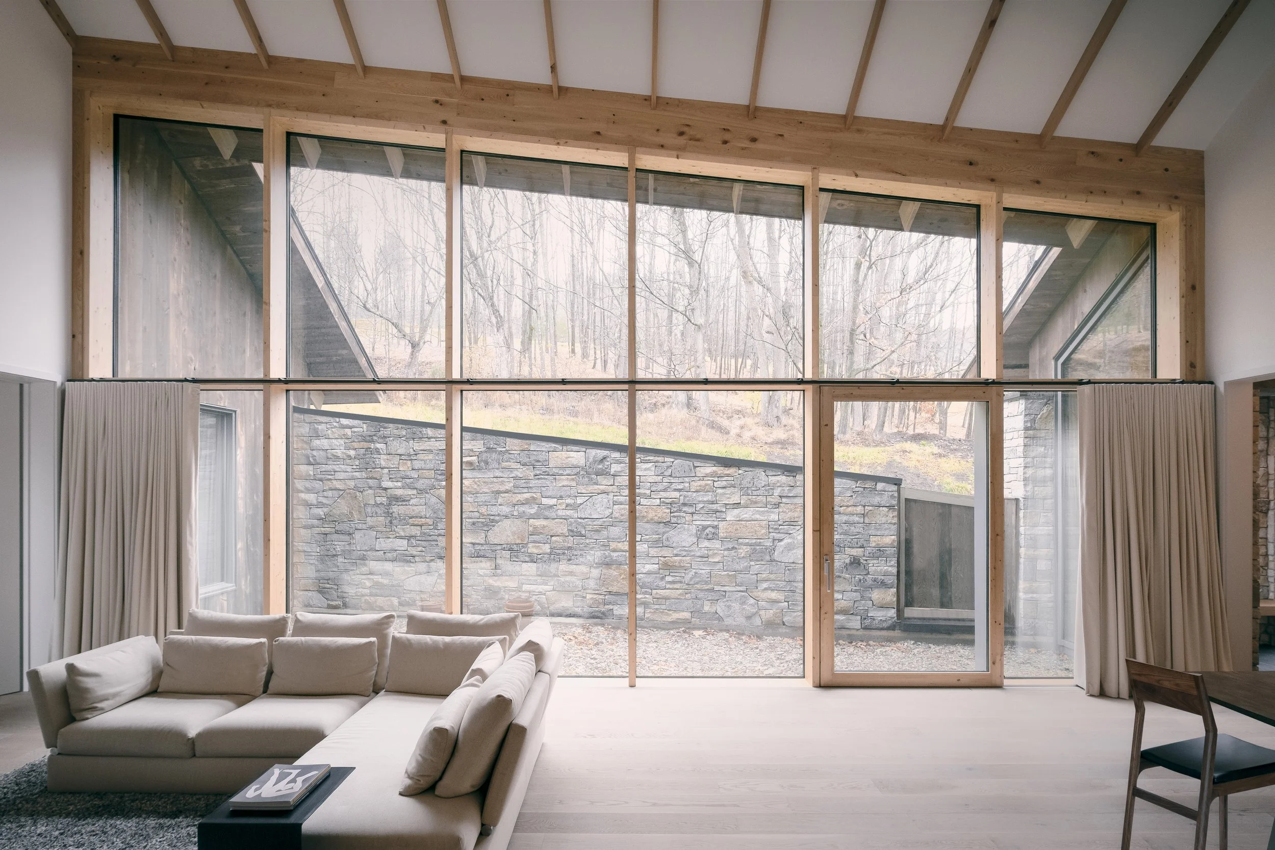 Living room with beige sectional sofa, black coffee table, wooden ceiling, large floor-to-ceiling windows with curtains, stone wall outside, and overcast trees in the background.