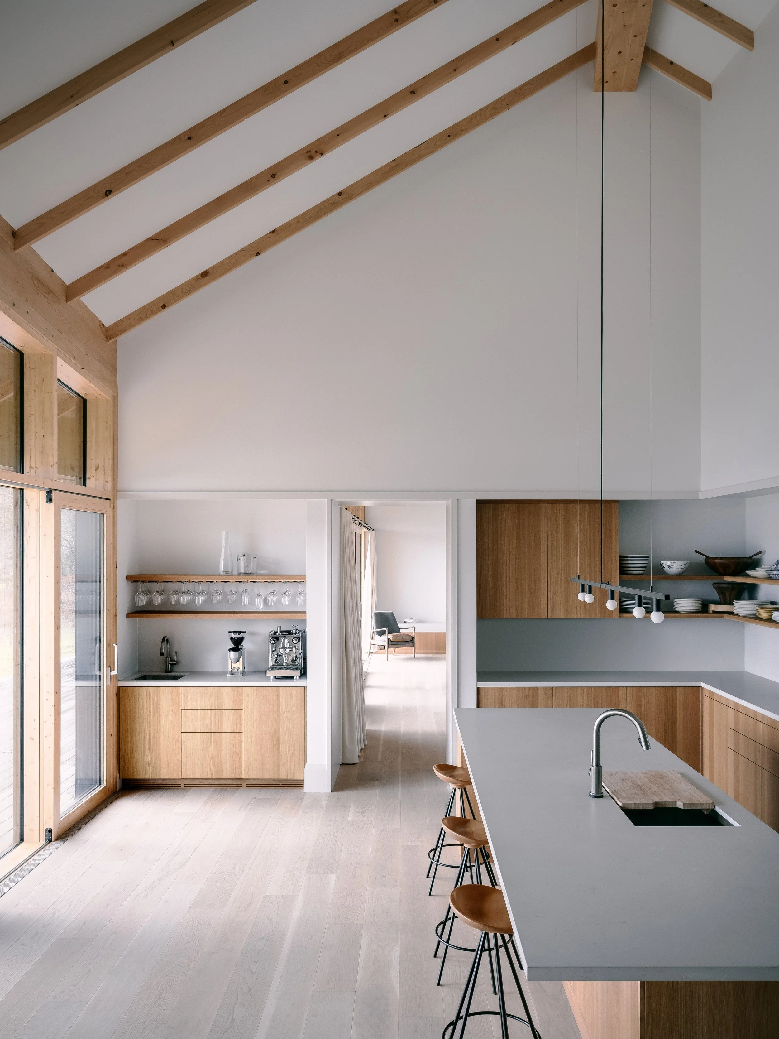Modern kitchen with wooden accents, white countertops, bar stools, and an open layout leading to a living area.