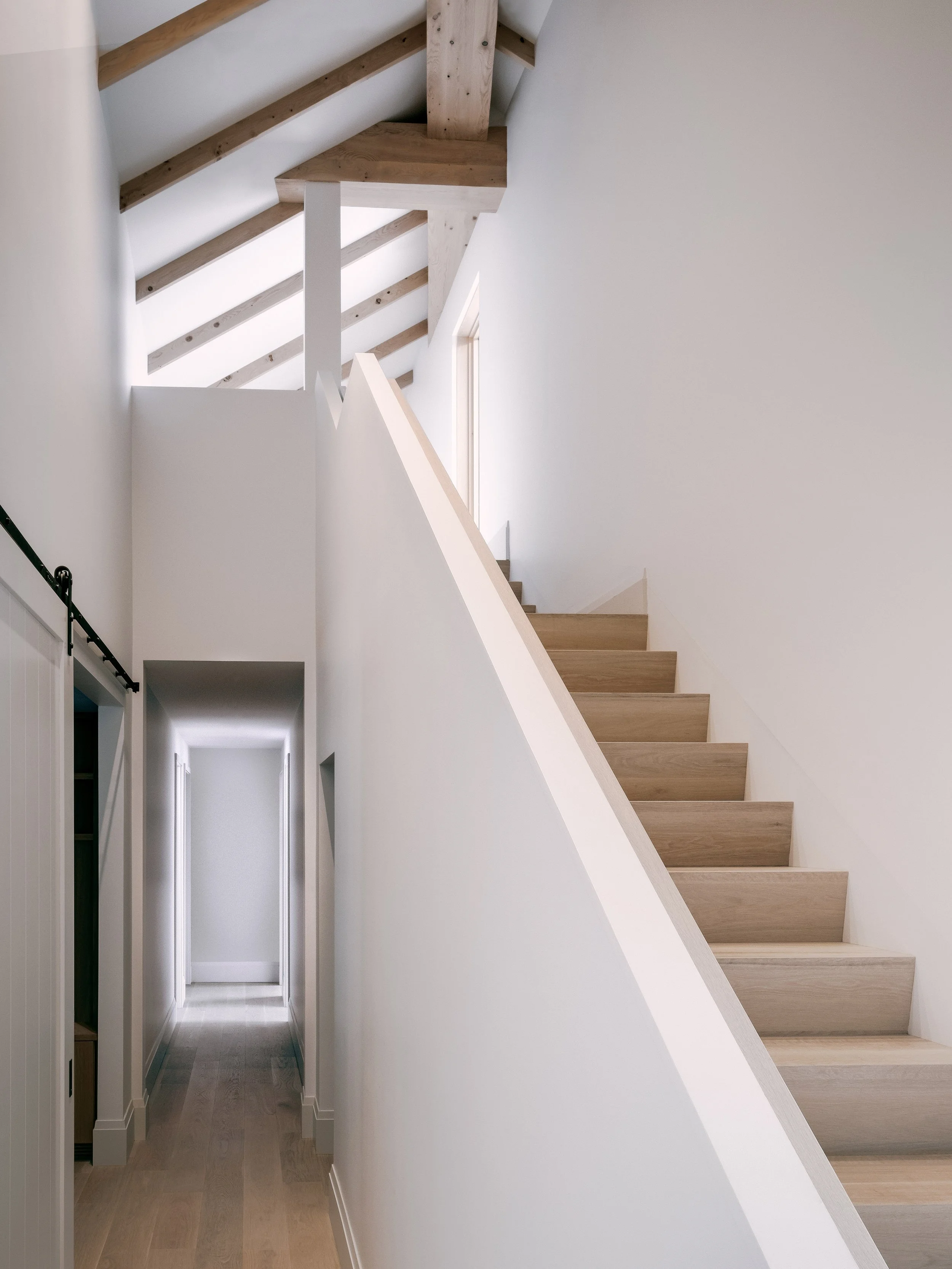 Interior view of a modern house with a white staircase and natural wood accents, including ceiling beams and stair treads, with a hallway at the bottom.