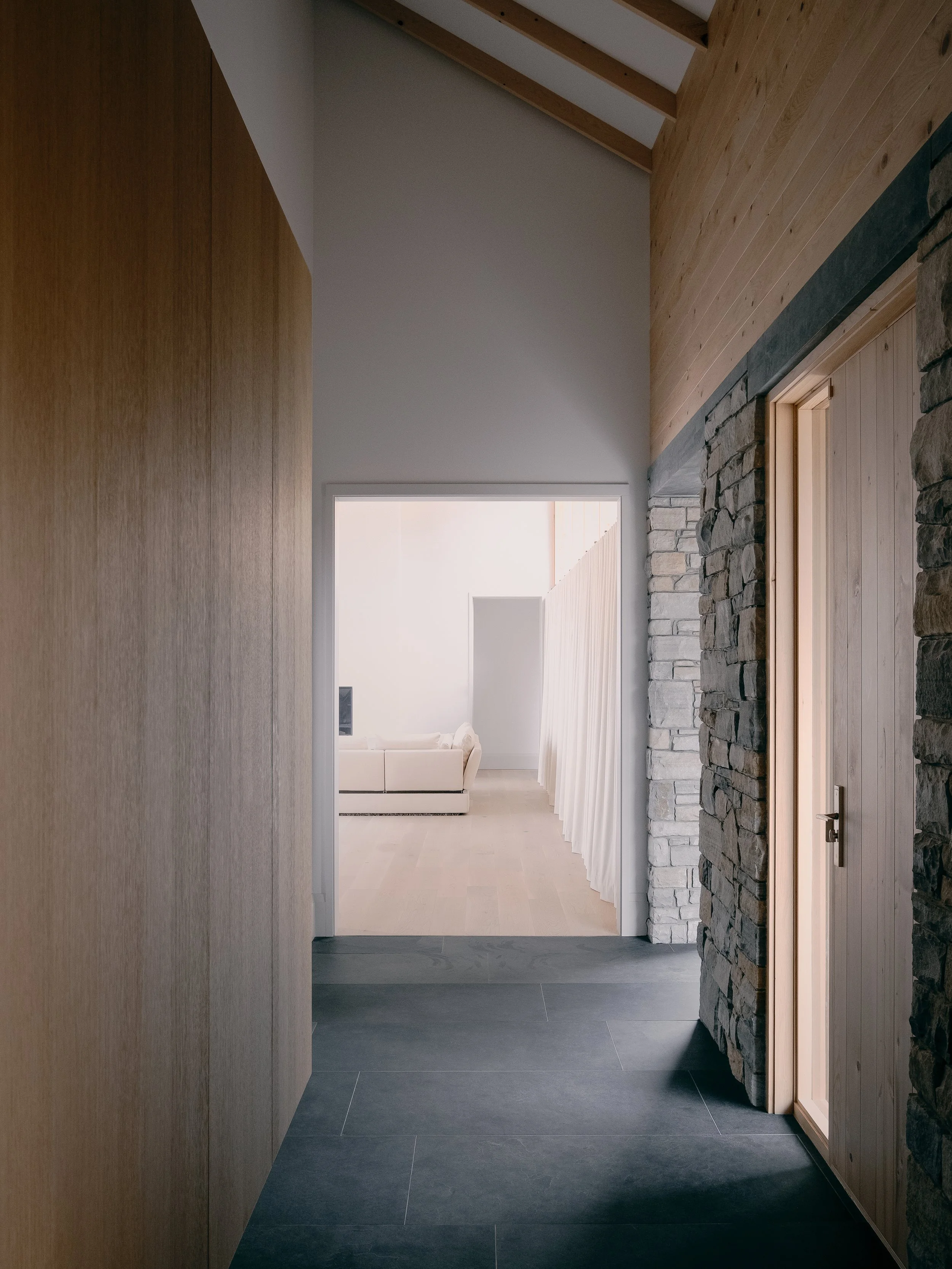 Interior hallway in a modern house with stone and wood walls, dark floor tiles, and a view into a bright living room with a white couch