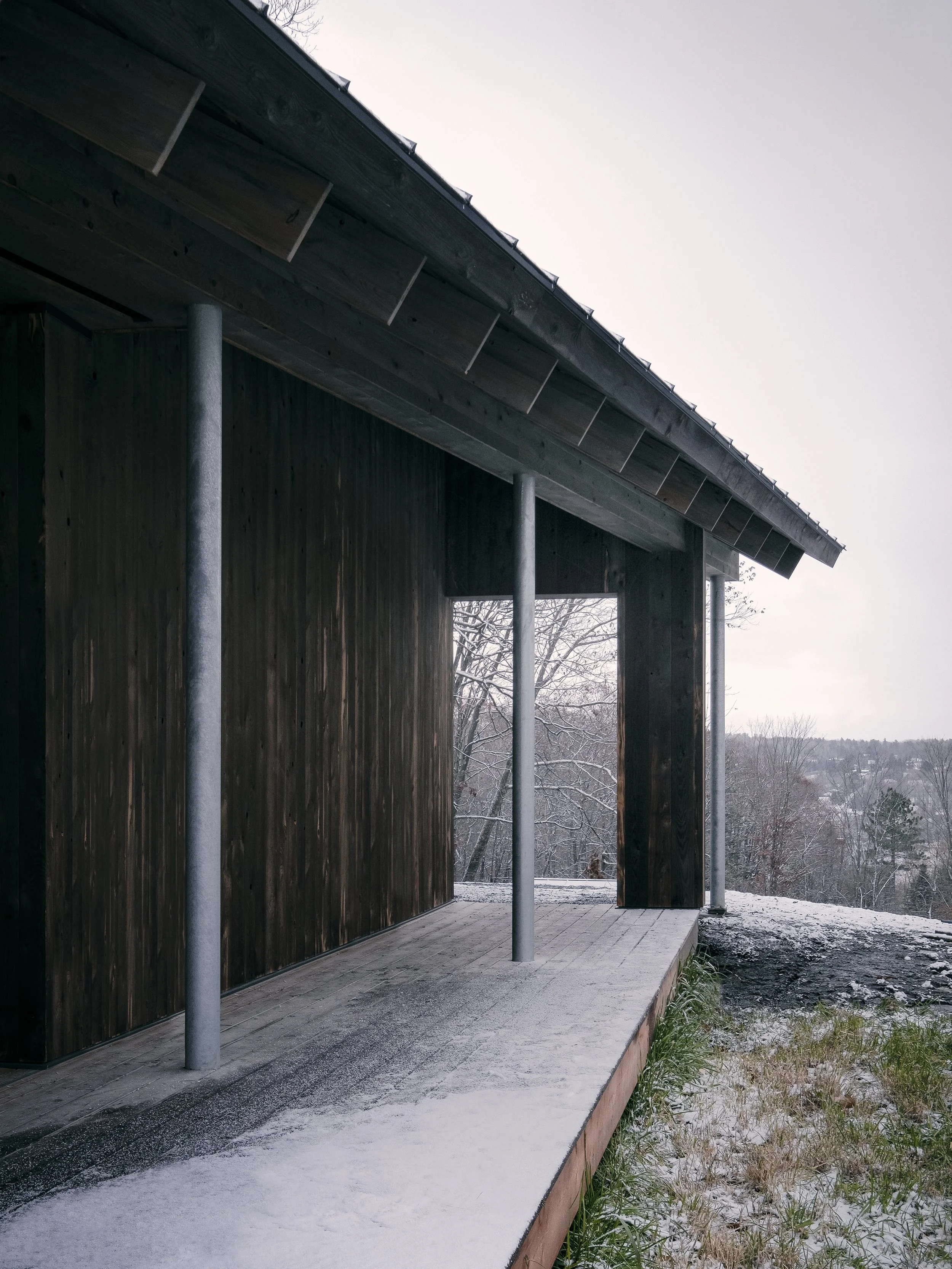 Side view of a rustic wooden porch with metal support posts, overlooking a snow-covered landscape with sparse trees.