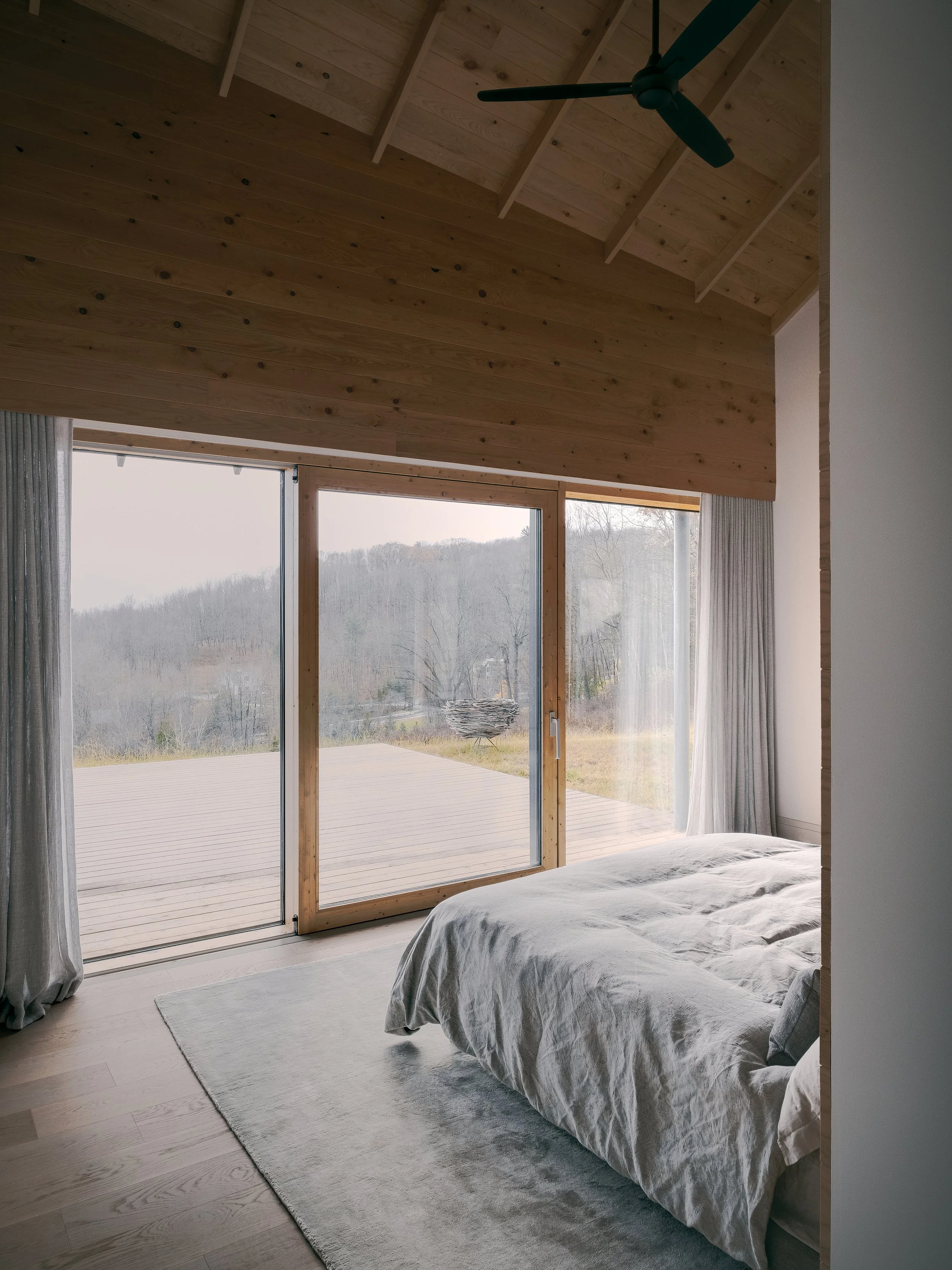 A bedroom with large floor-to-ceiling sliding glass doors, view of a deck and a rural landscape beyond. The room has a wooden ceiling with a ceiling fan, light-colored wooden floor, and a bed with light-colored bedding.
