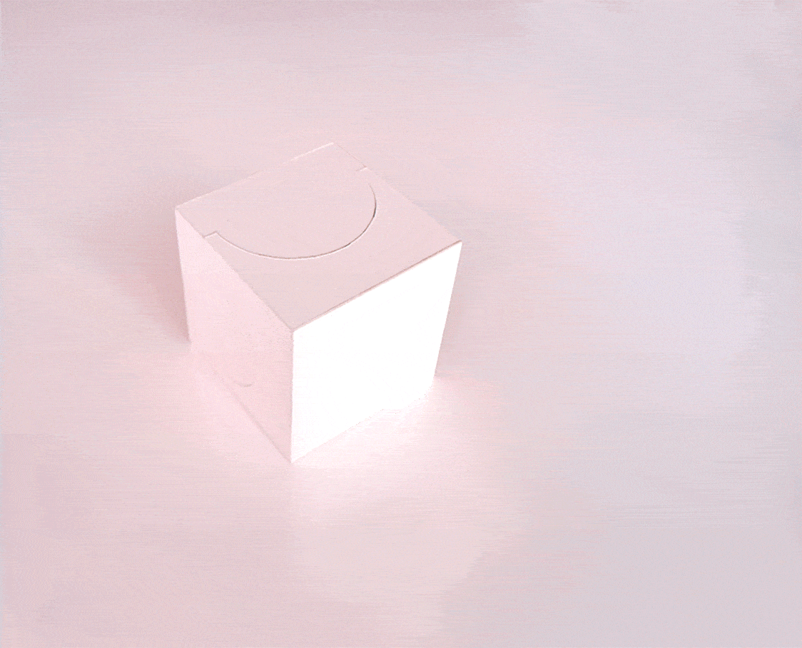 A white cube with a semi-circular cutout on the top face, placed on a pink surface.