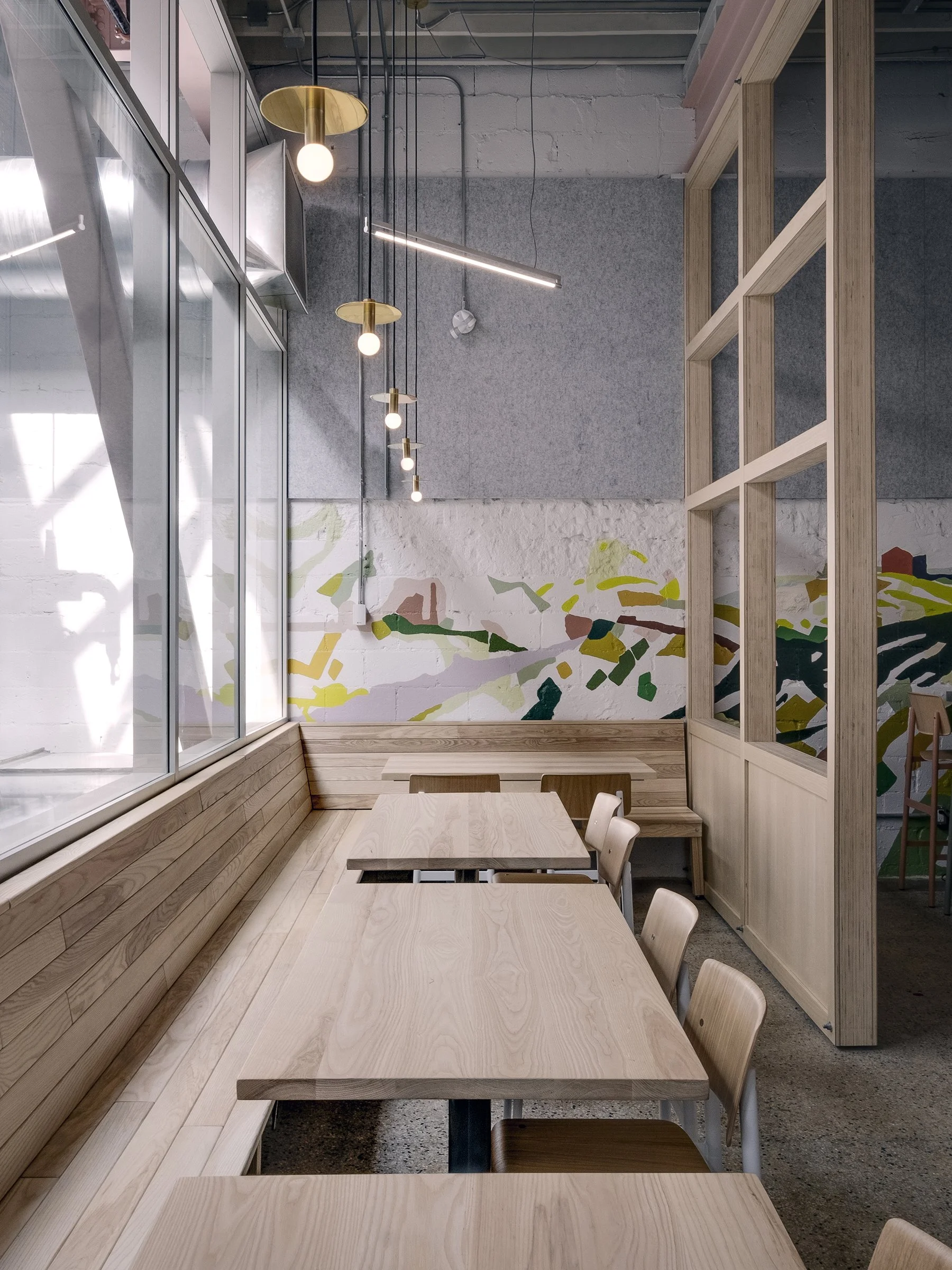 Empty modern cafe with wooden tables, chairs, and bench seating by large window, decorative lighting fixtures, and a painted mural on the wall.