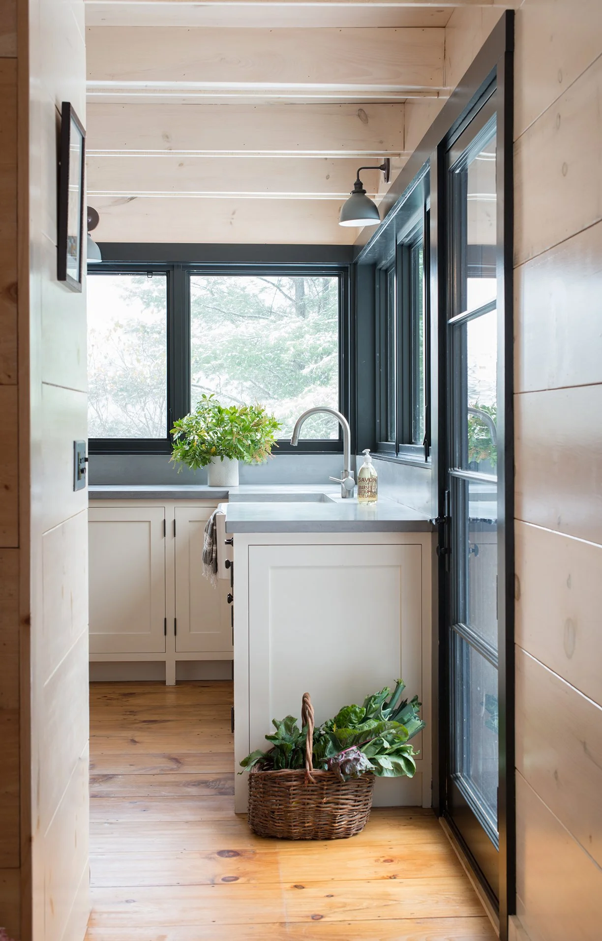 A cozy kitchen with natural wood walls and ceiling, black-framed windows, a white cabinet, a sink with a silver faucet, a potted plant on the counter, a wire basket filled with leafy greens on the floor, and natural light streaming through to illuminate the space.