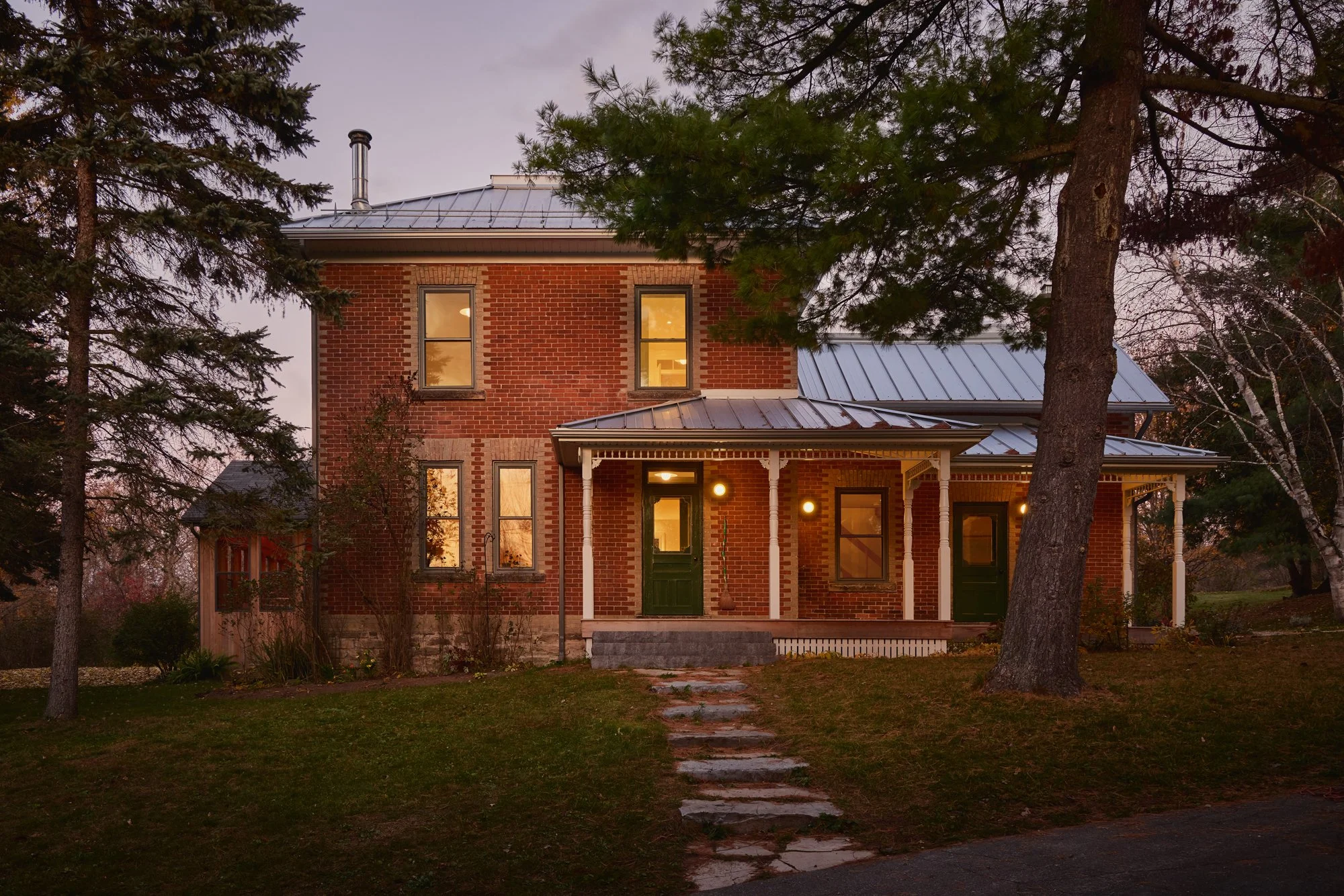 A two-story brick house with a metal roof and front porch, surrounded by trees at dusk