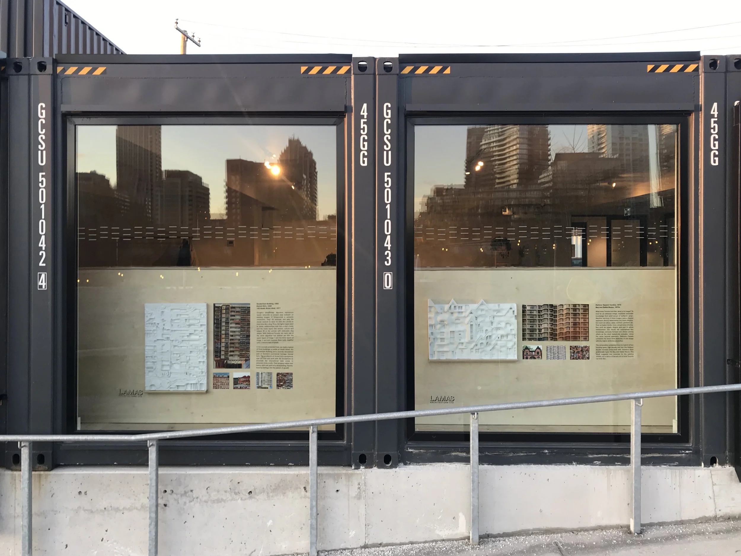 Two display cases containing architectural models and information panels, with tall city buildings reflected in the glass and a construction barrier in the foreground.