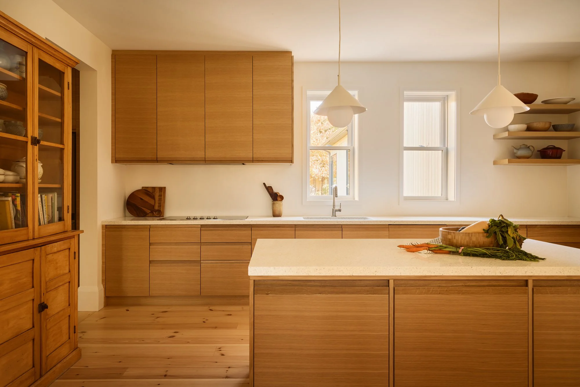 Minimalist kitchen with wooden cabinets, shelves, and flooring, white countertops, two pendant lights, and two windows with autumn foliage outside.