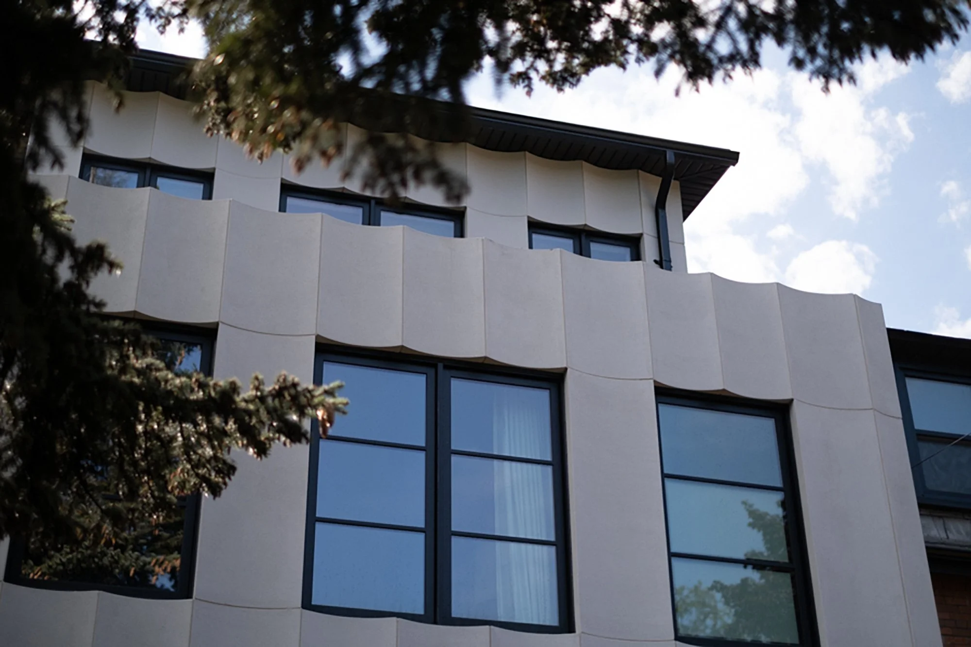 Close-up of modern building with wavy beige facade and large rectangular windows, partly obscured by tree branches.