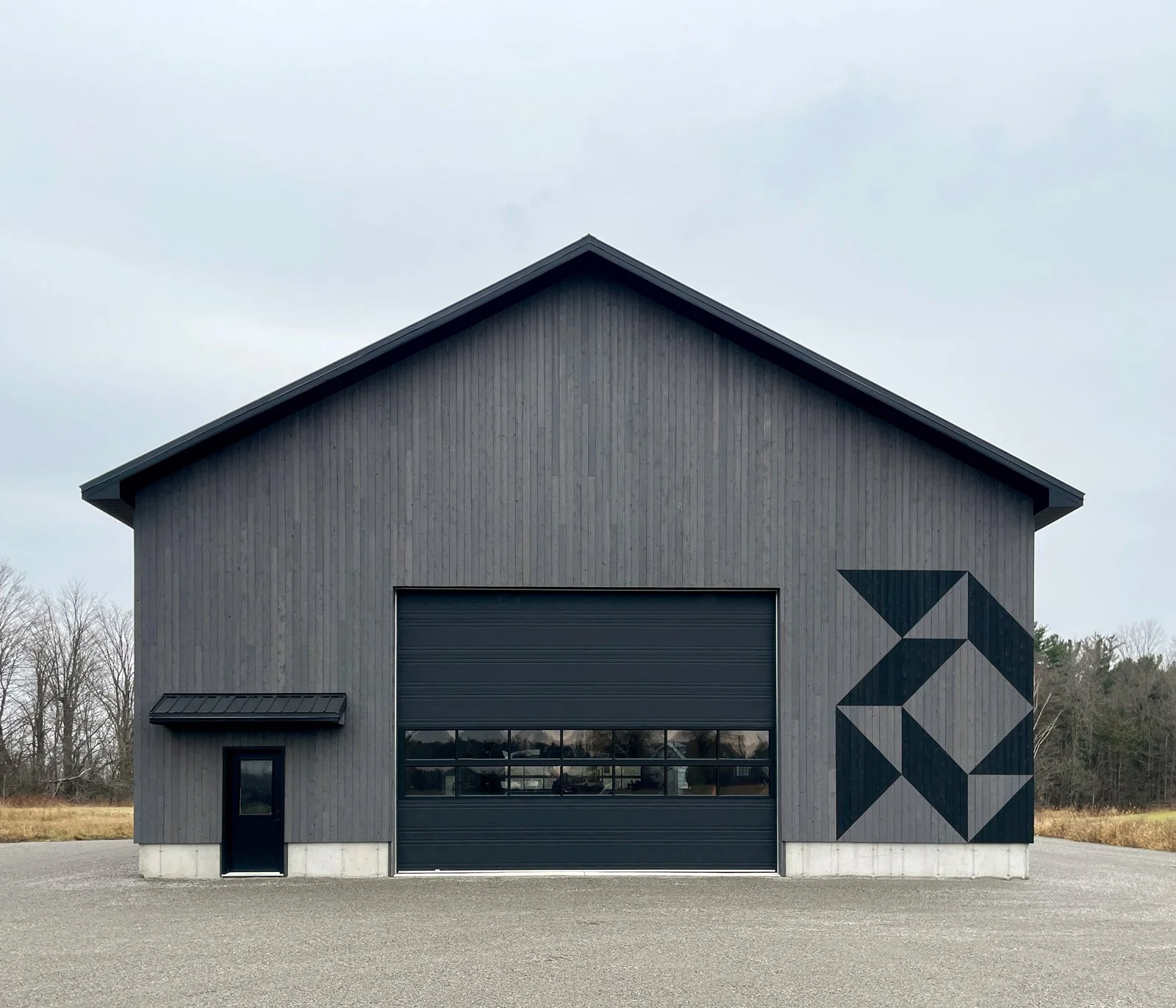 A modern gray barn-style building with a large garage door, a side door with a small roof, and a geometric black mural on the right side wall, set against a cloudy sky and surrounded by leafless trees.