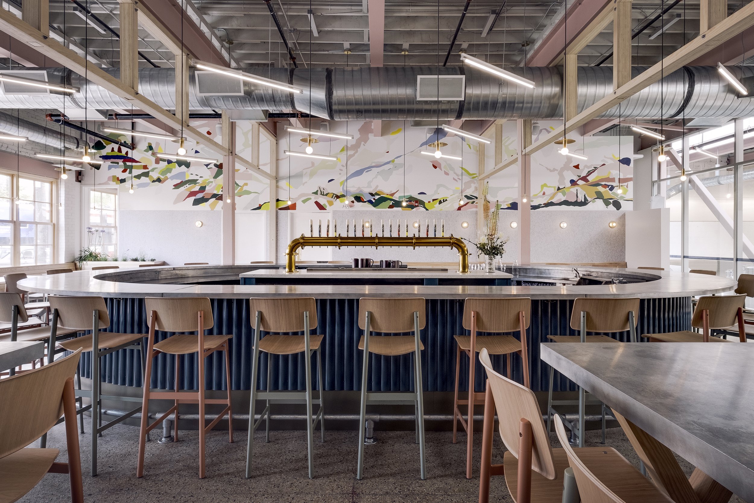 Modern restaurant interior with a circular bar, wooden chairs, colorful wall art, and industrial ceiling ductwork.