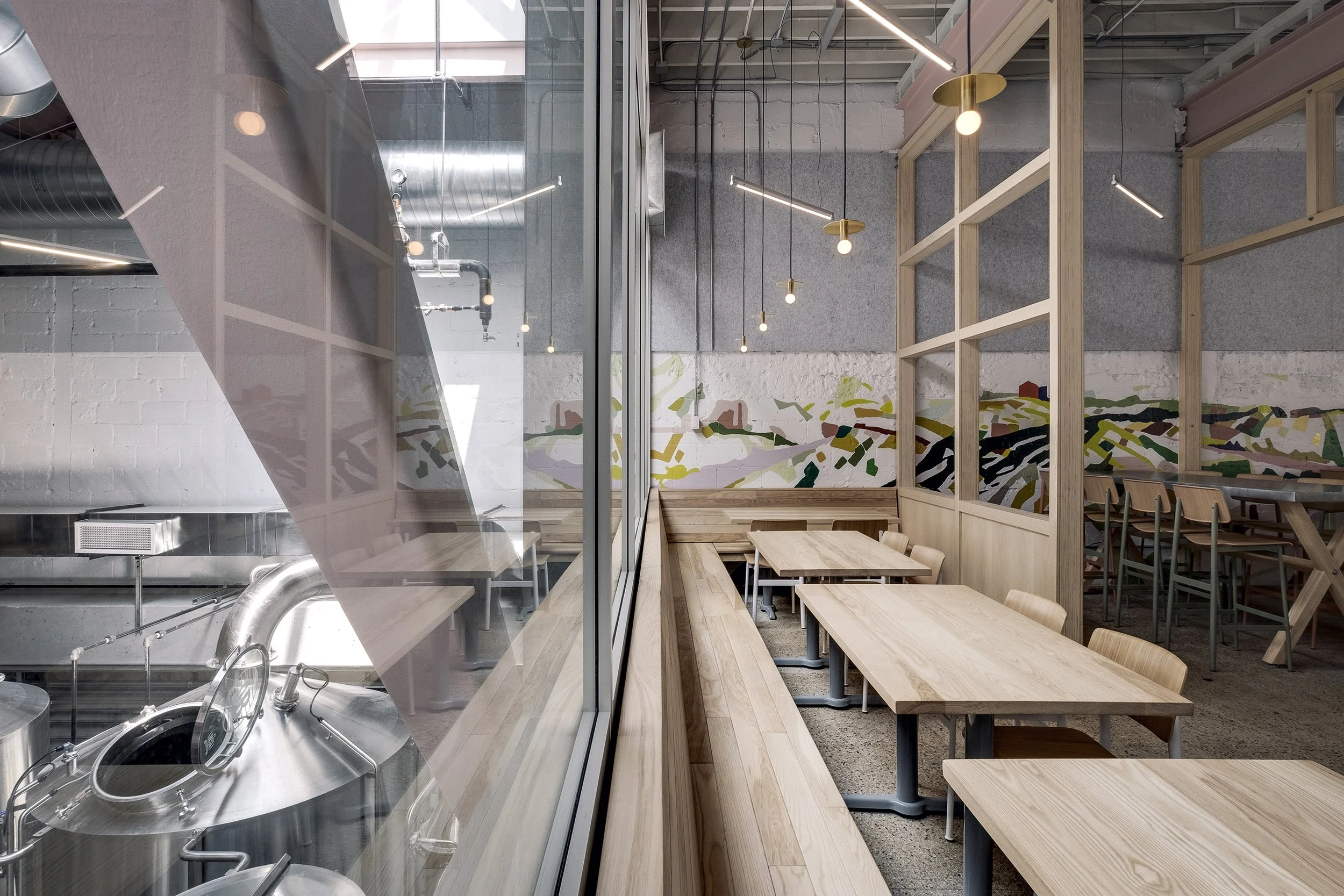 Interior of a modern restaurant or cafe with wooden tables and chairs, pendant lights, a painted mural on the wall, and a glass partition separating the dining area from the kitchen.