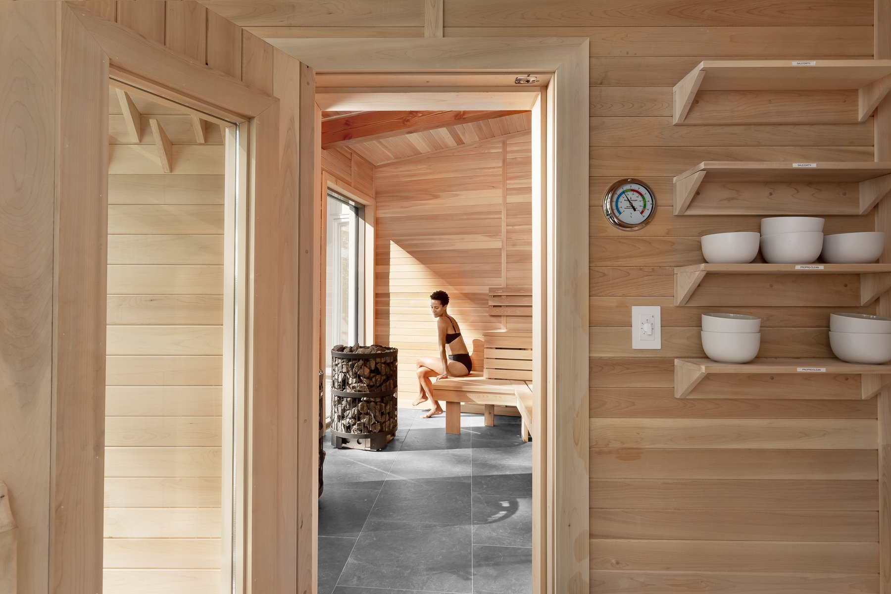 A woman in a sauna room with wooden walls, sitting on a bench next to a woodpile heater exposed to sunlight.