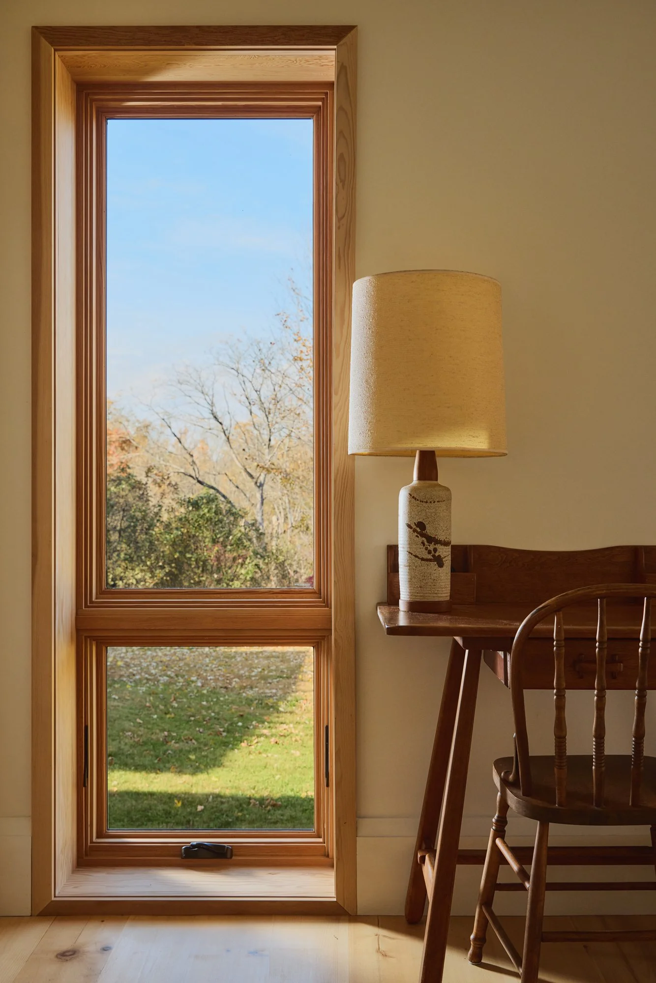 A room corner with a wooden framed glass door, a wooden chair, a wooden desk with a ceramic table lamp, and a view of trees and grass outside during daytime.
