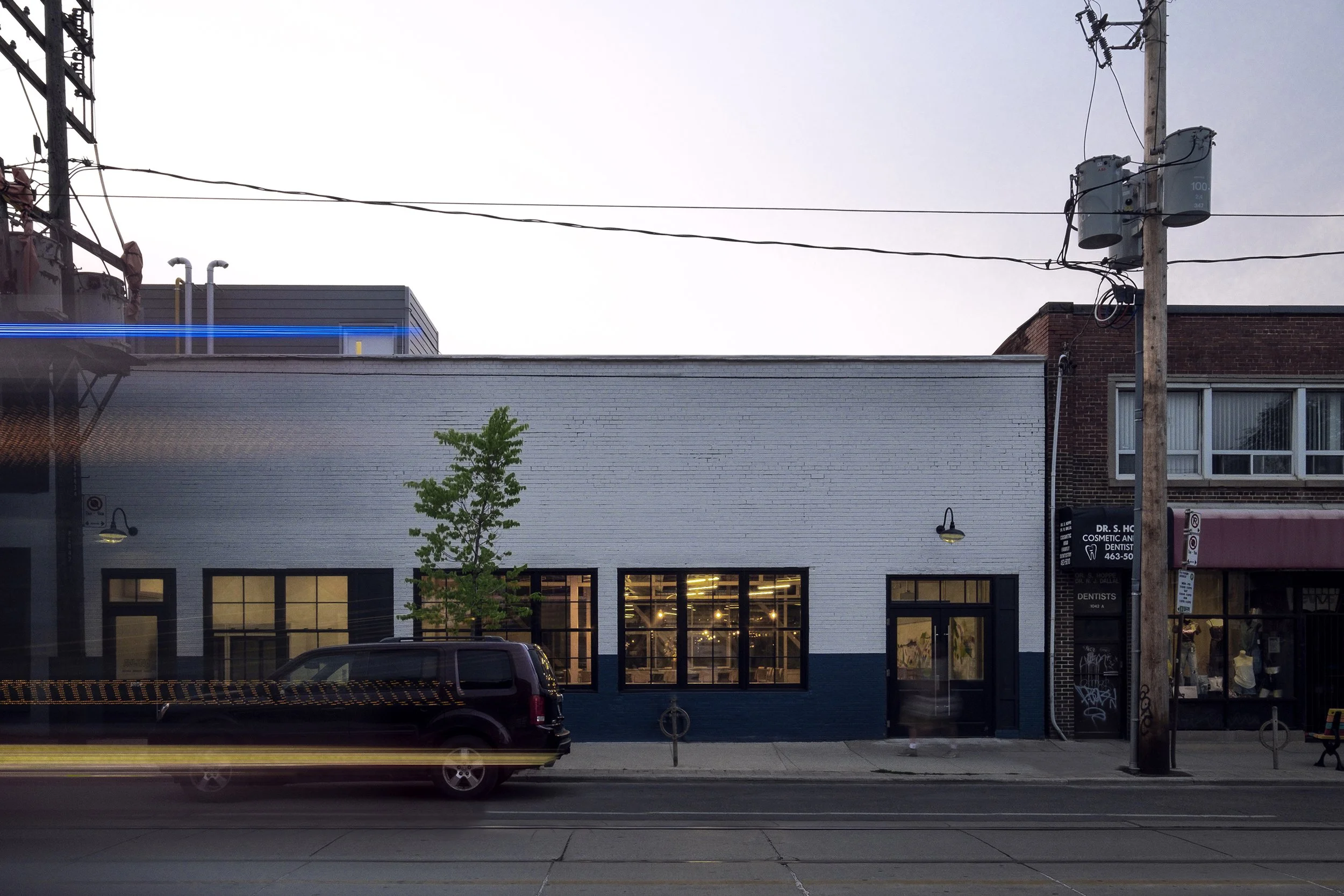 Street scene with a white building, two large windows, a small tree, and a parked black SUV, with a utility pole and overhead wires.