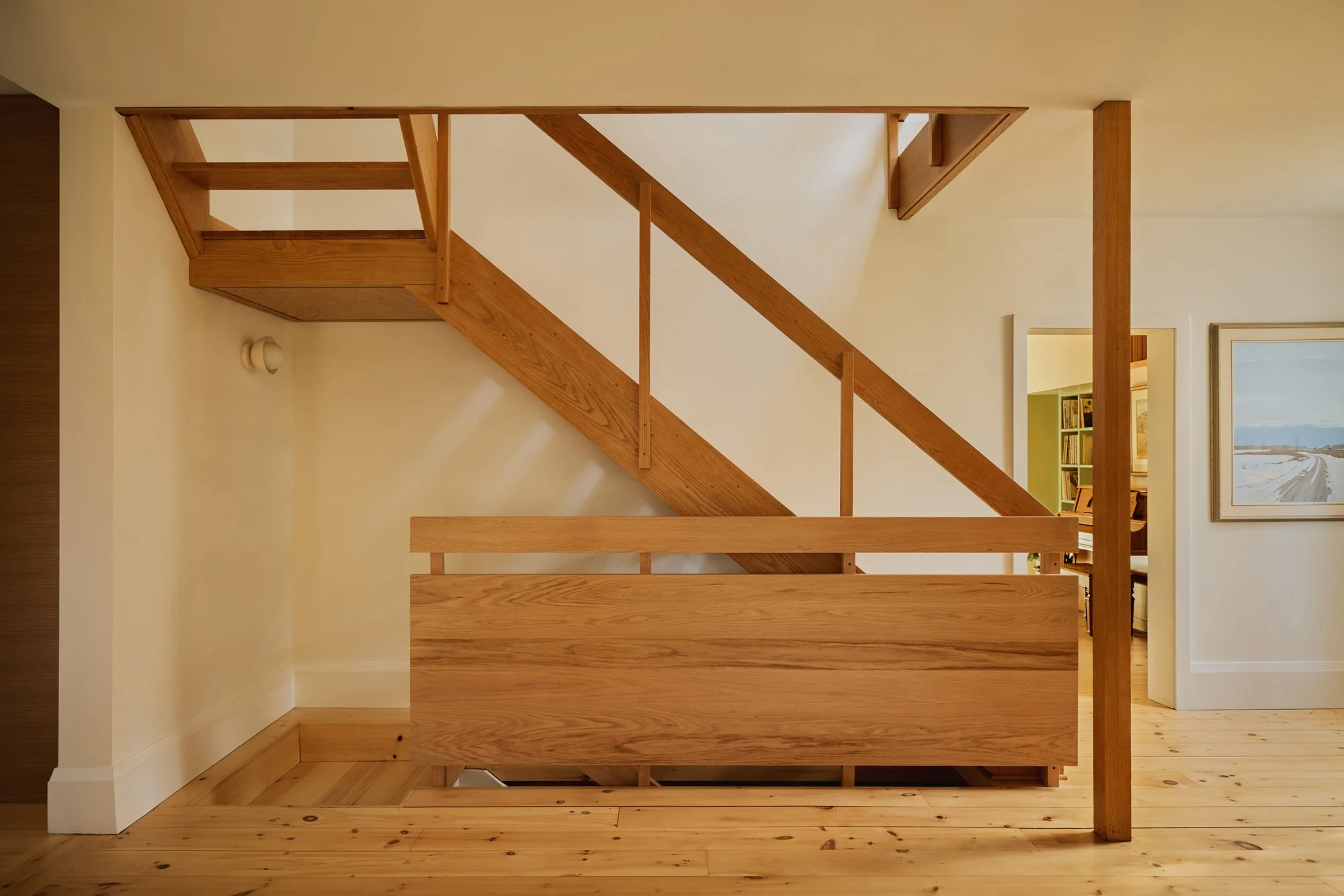 Interior view of a wooden staircase with railing in a home, visible next to a doorway leading to a room with a bookshelf and a painting on the wall.