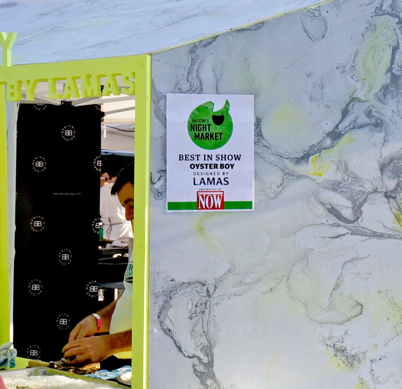 A sign on a marble wall advertising a night market oyster show, with a person preparing oysters visible through a green-framed window.