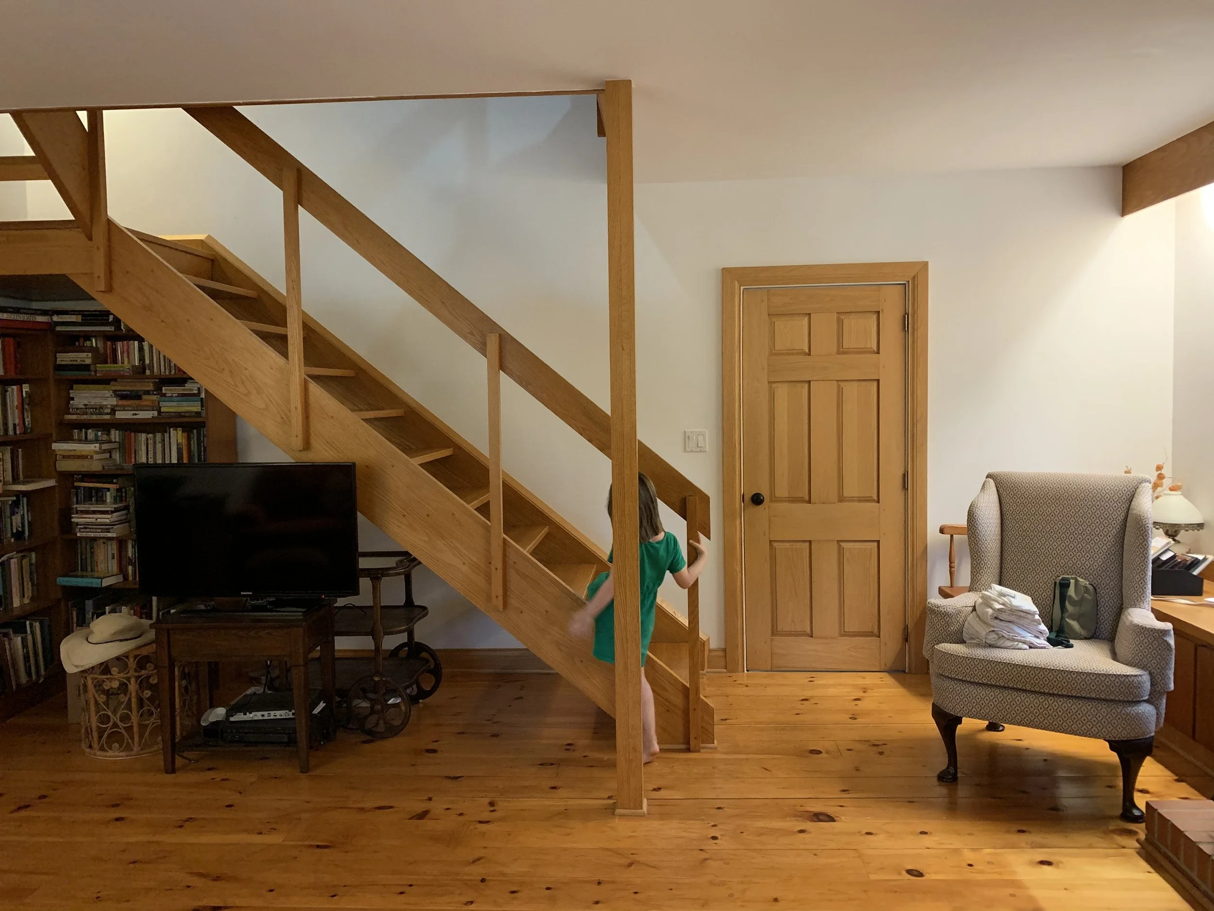 Living room with wooden staircase, bookshelf, flat-screen TV, armchair, and a child in a green shirt climbing the stairs.