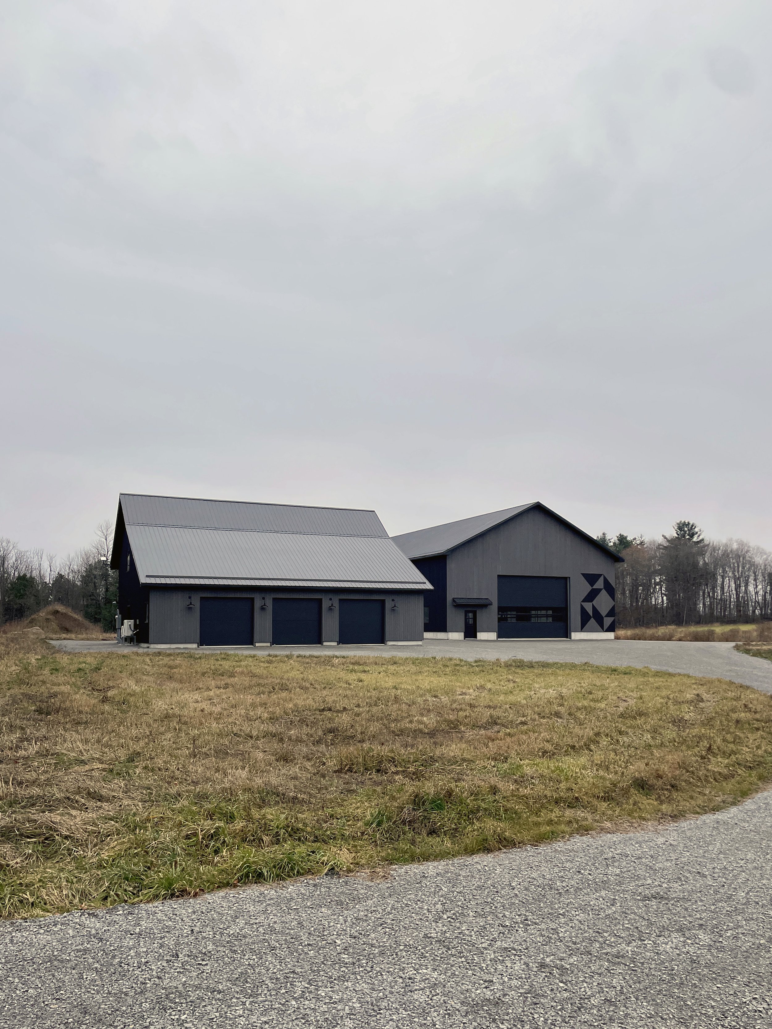 Modern gray building with three garage doors and a second garage entrance, surrounded by a gravel driveway and a grassy yard, with trees in the distance under an overcast sky.