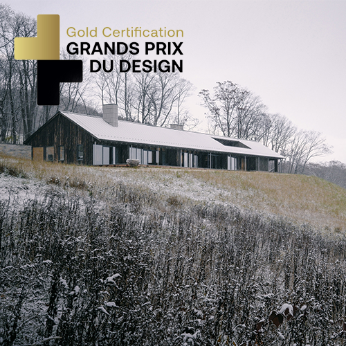 A house in a snowy field with leafless trees in the background, overlaid with text that reads 'Gold Certification GRANDS PRIX DU DESIGN' and stylized plus signs.