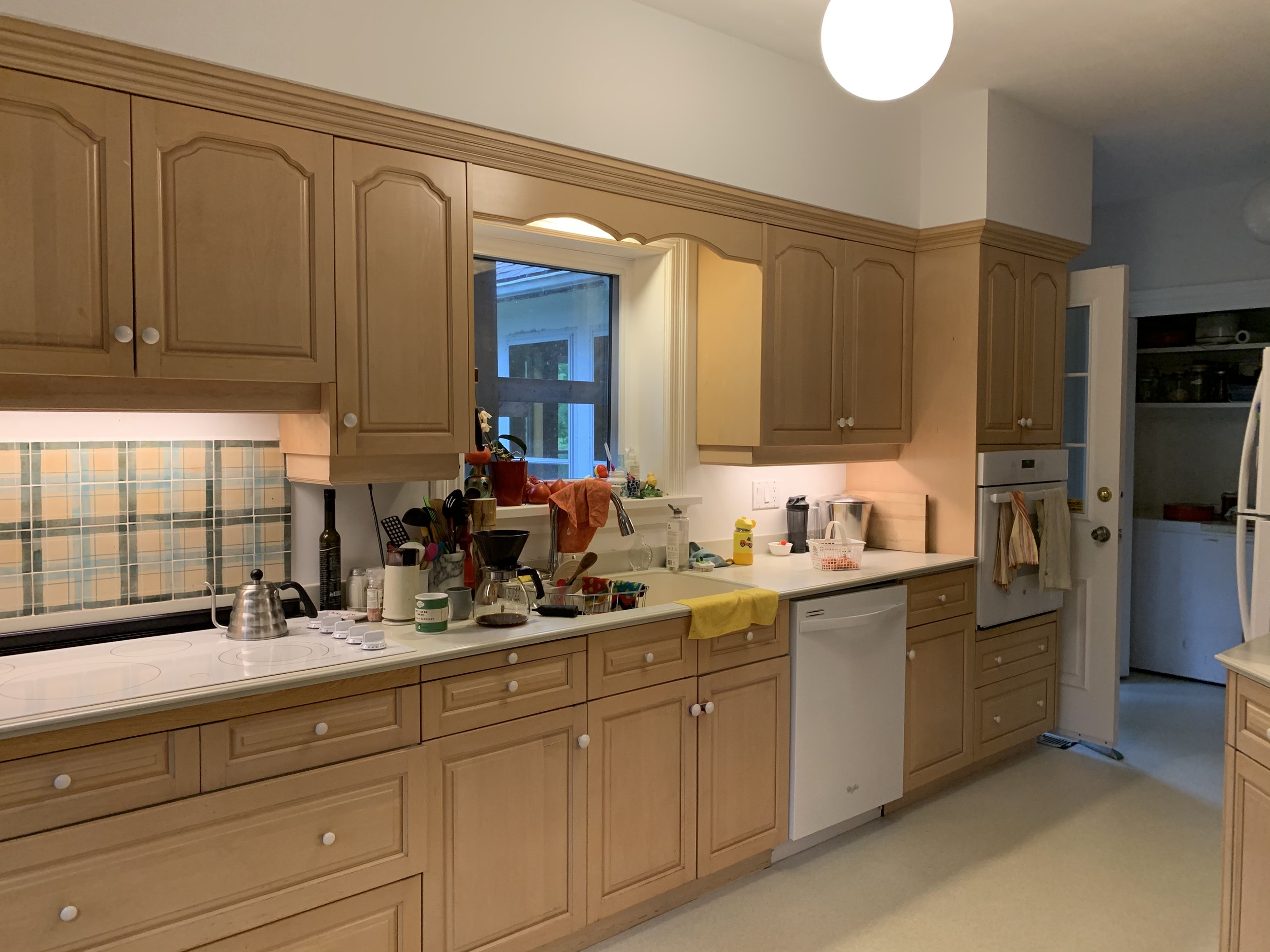 A kitchen with wooden cabinets, a window above the sink, and various kitchen items on the countertop including a coffee maker, utensils, and a yellow towel. There's a dishwasher below the counter, and a doorway leading to another room.