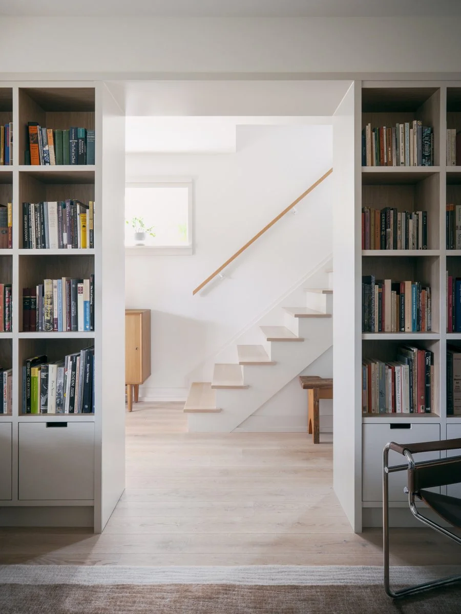 A bright interior view showing white bookshelves filled with books on each side of an open doorway, leading to a staircase with white steps and a wooden handrail, and a small window letting in natural light.