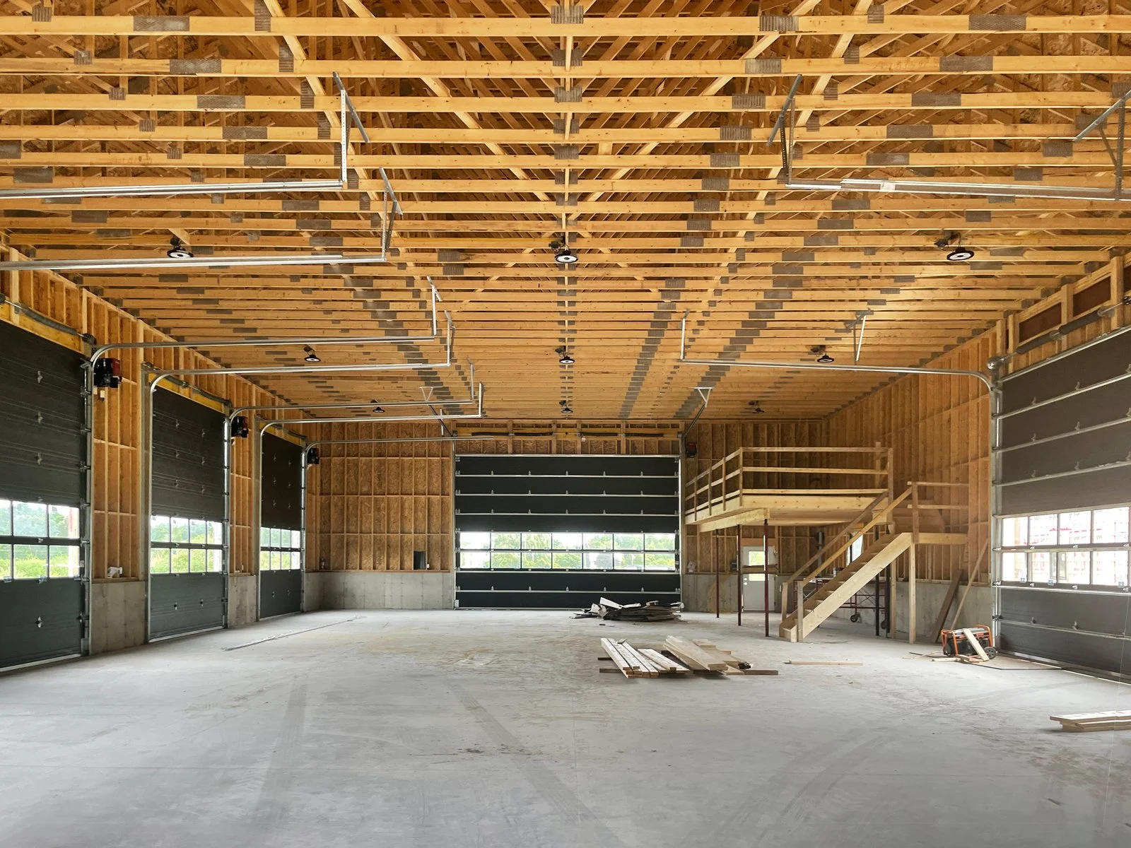 Interior of a building under construction with exposed wooden walls and ceiling, large garage-style doors, and a wooden staircase leading to an upper platform.