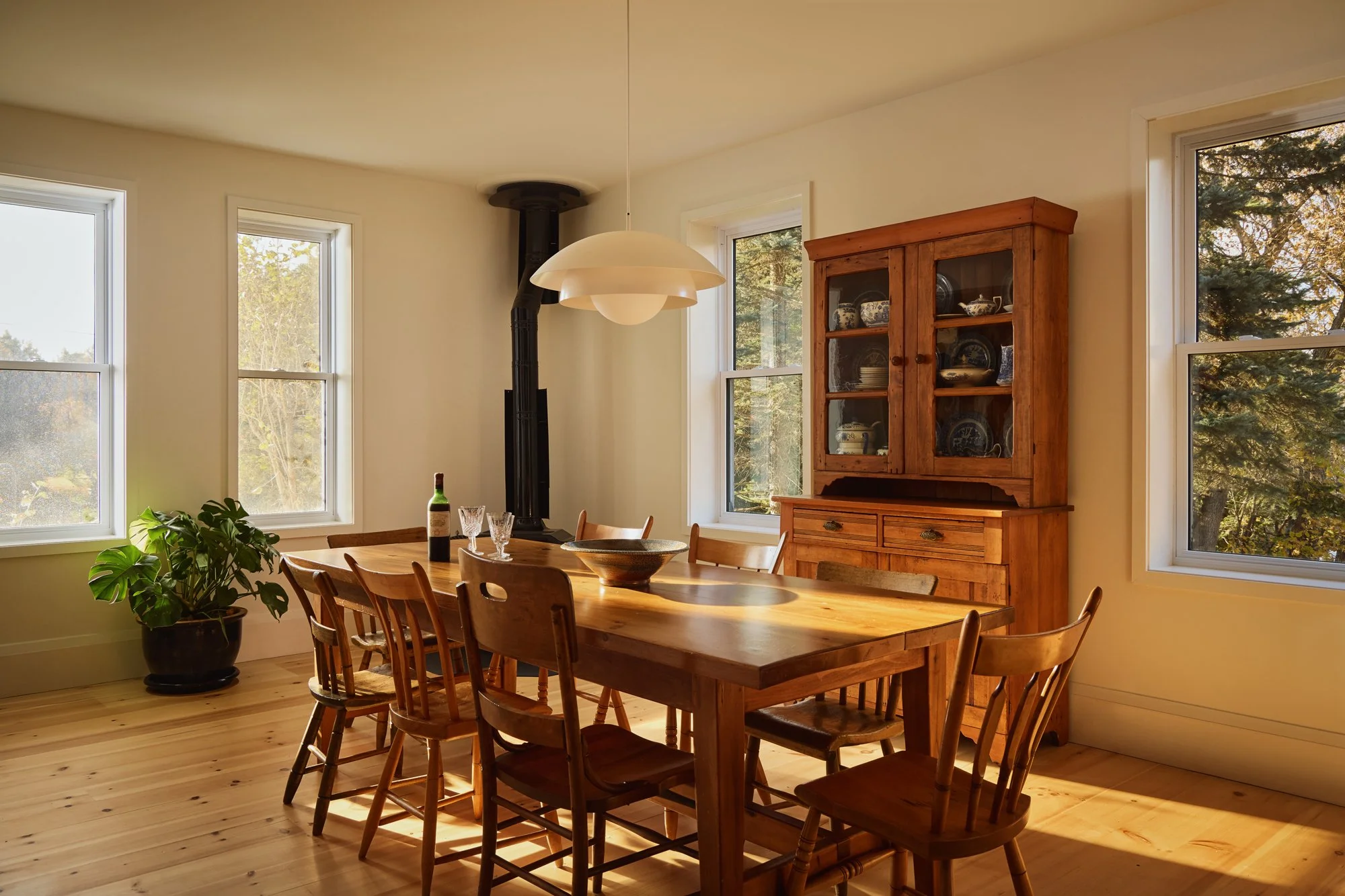 A wooden dining table with eight matching chairs, a potted plant in the corner, a glass bottle with wine and two wine glasses on the table, a wooden hutch with dishes, three windows letting in natural light, and a black wood stove in the corner.