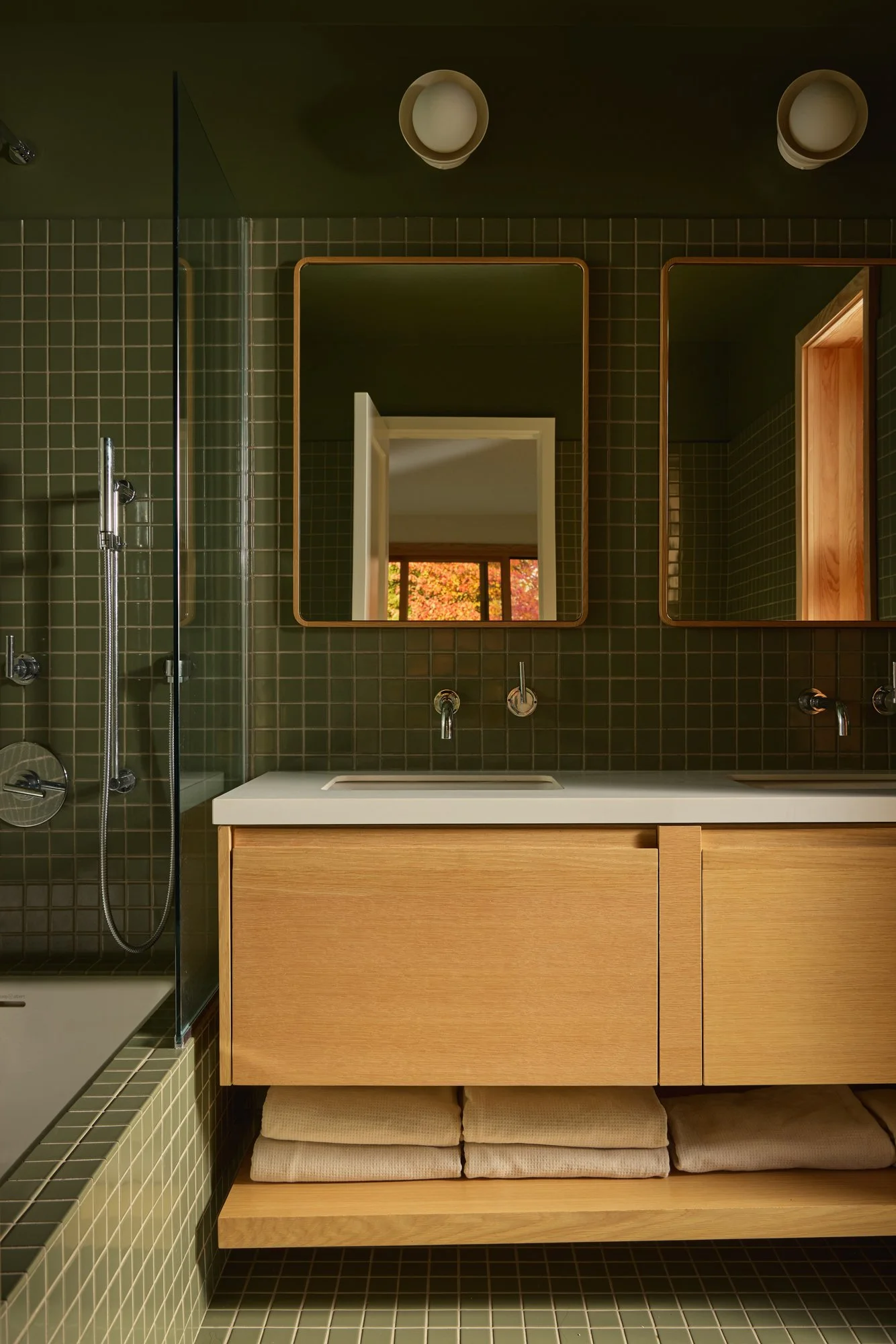 Modern bathroom with green tiled walls and floor, a wooden vanity with a white countertop, two mirrors, and two wall-mounted faucets. There is a shower area with a glass partition and a window showing autumn foliage outside.