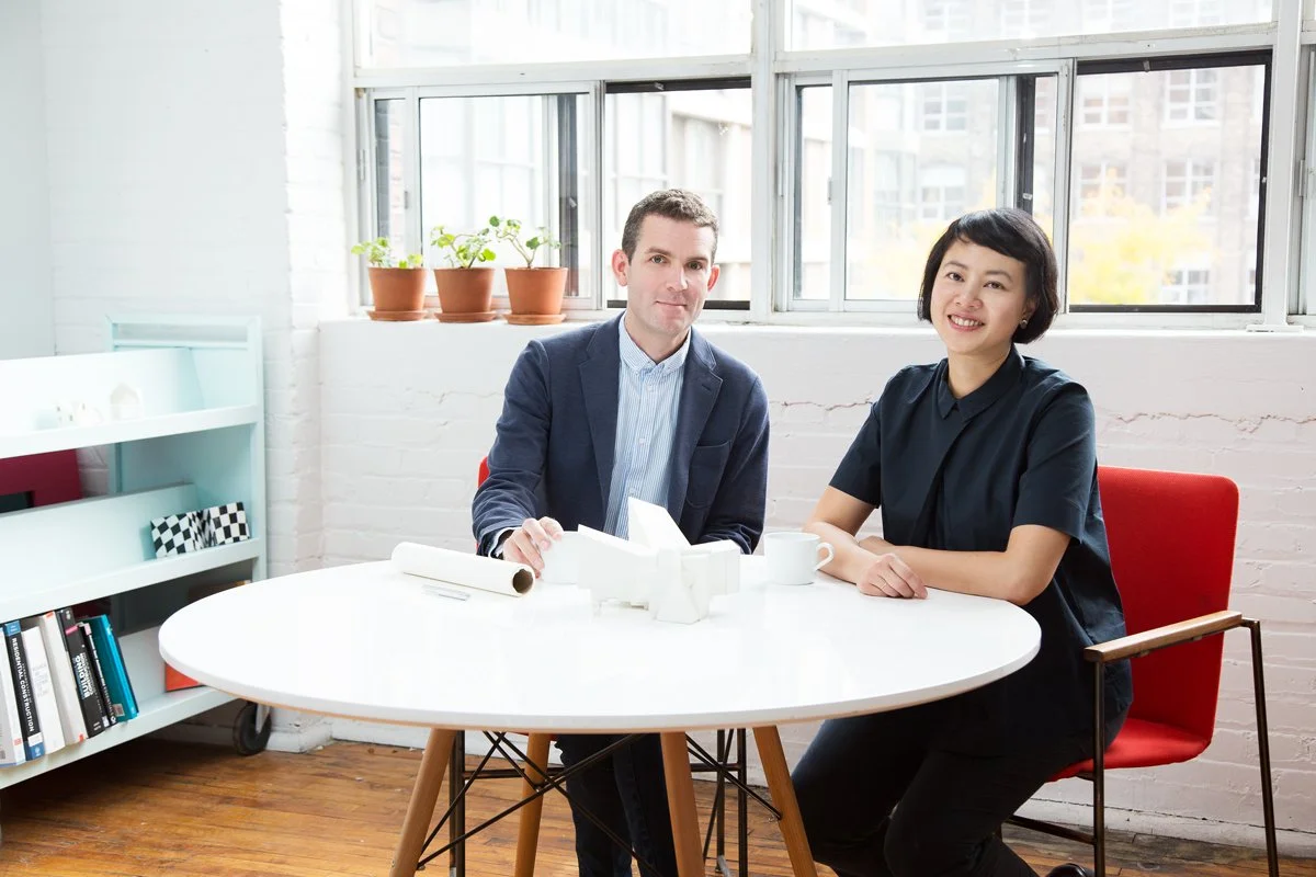 Two people sitting at a round white table in a bright room with large windows; a man in a suit and a woman with short black hair smiling at the camera; potted plants on the windowsill.