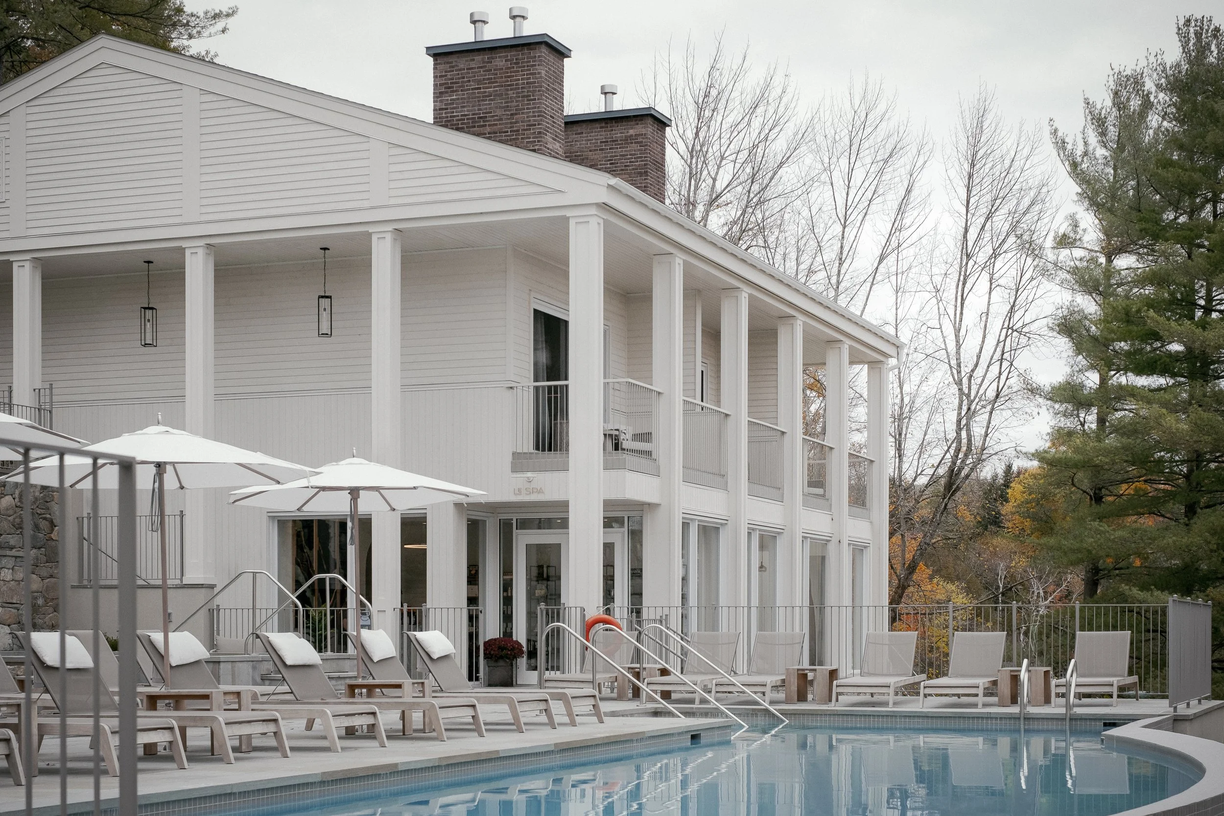 A pool area outside a white building with a balcony, lounge chairs, umbrellas, and trees in the background.
