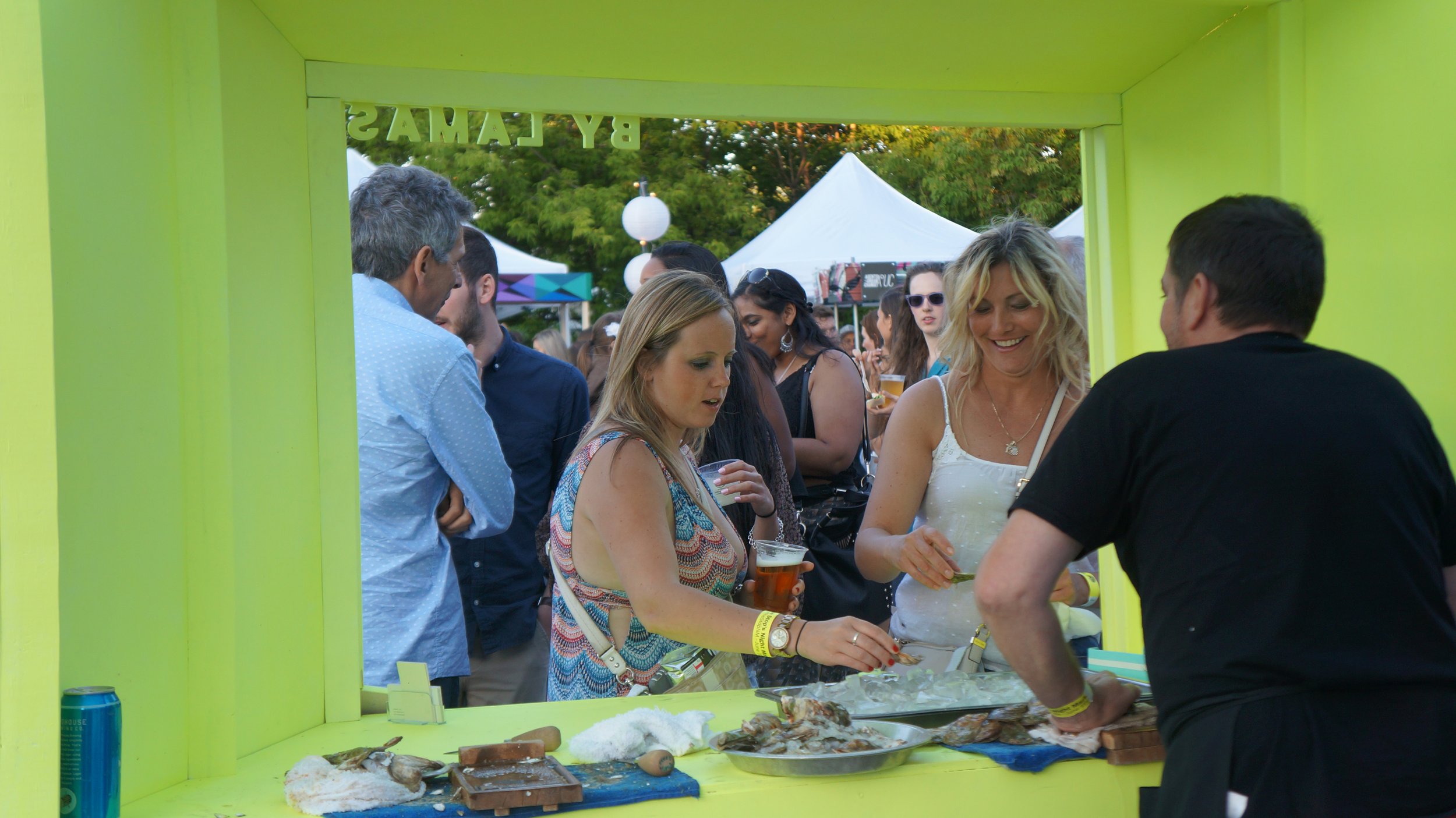 People gathered at a bright yellow food stand at an outdoor festival, with tents and trees in the background, and food preparation on the counter.