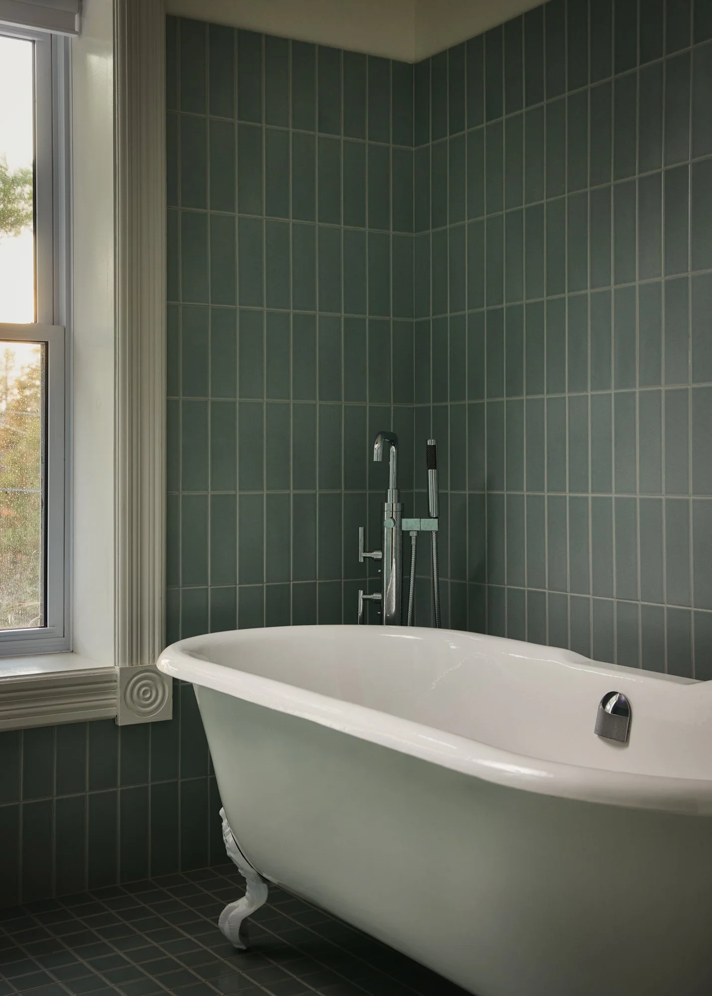 Vintage white clawfoot bathtub with green tiled wall and a window with a view of trees outside.