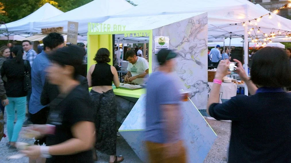 Crowd at an outdoor event with tents, including a booth called 'Oyster Day' serving oysters, and people taking photos and enjoying food.