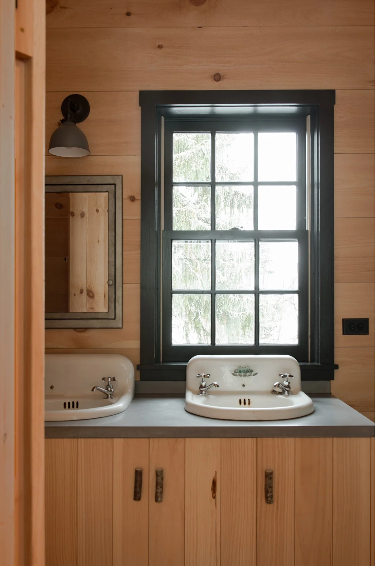 A rustic bathroom interior with two vintage sinks, a large window with black trim, wooden walls, and a wall-mounted light fixture.