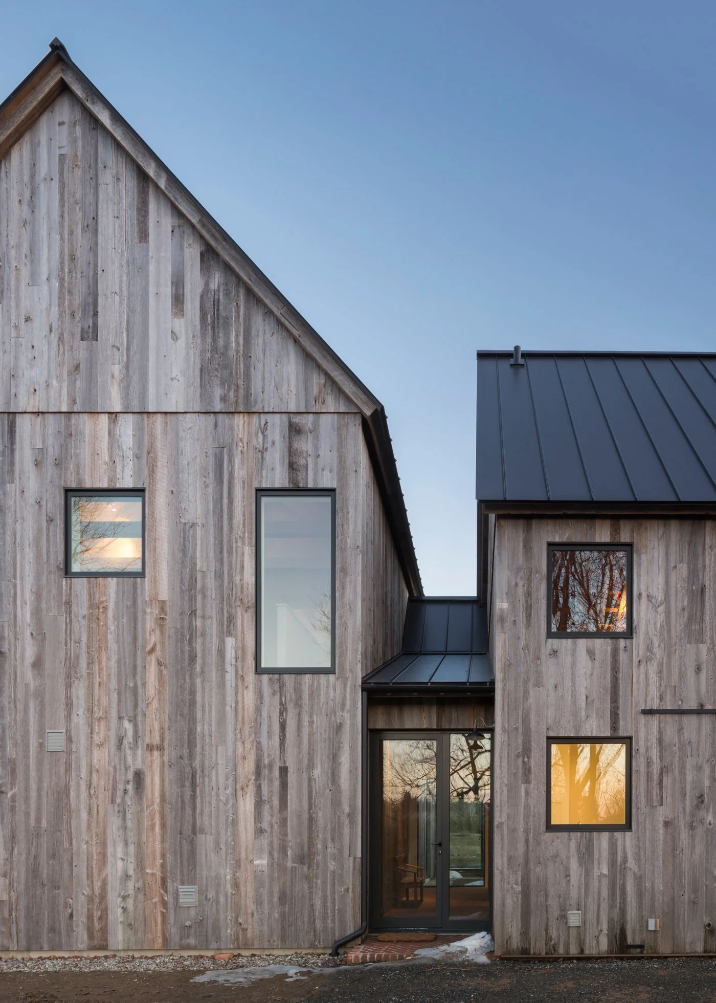 Front view of a modern barn-style house with weathered wooden exterior, black metal roof, and large windows at sunset.
