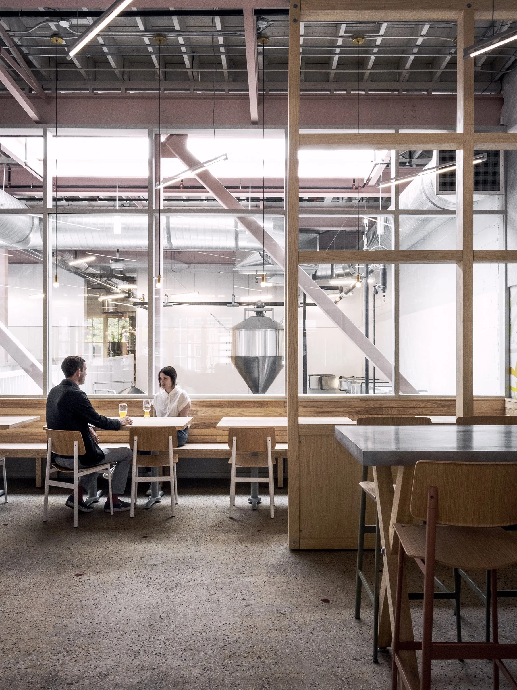 A man and woman sitting at a wooden table in a modern, industrial-style cafe, with large glass partitions showing brewing equipment in the background.